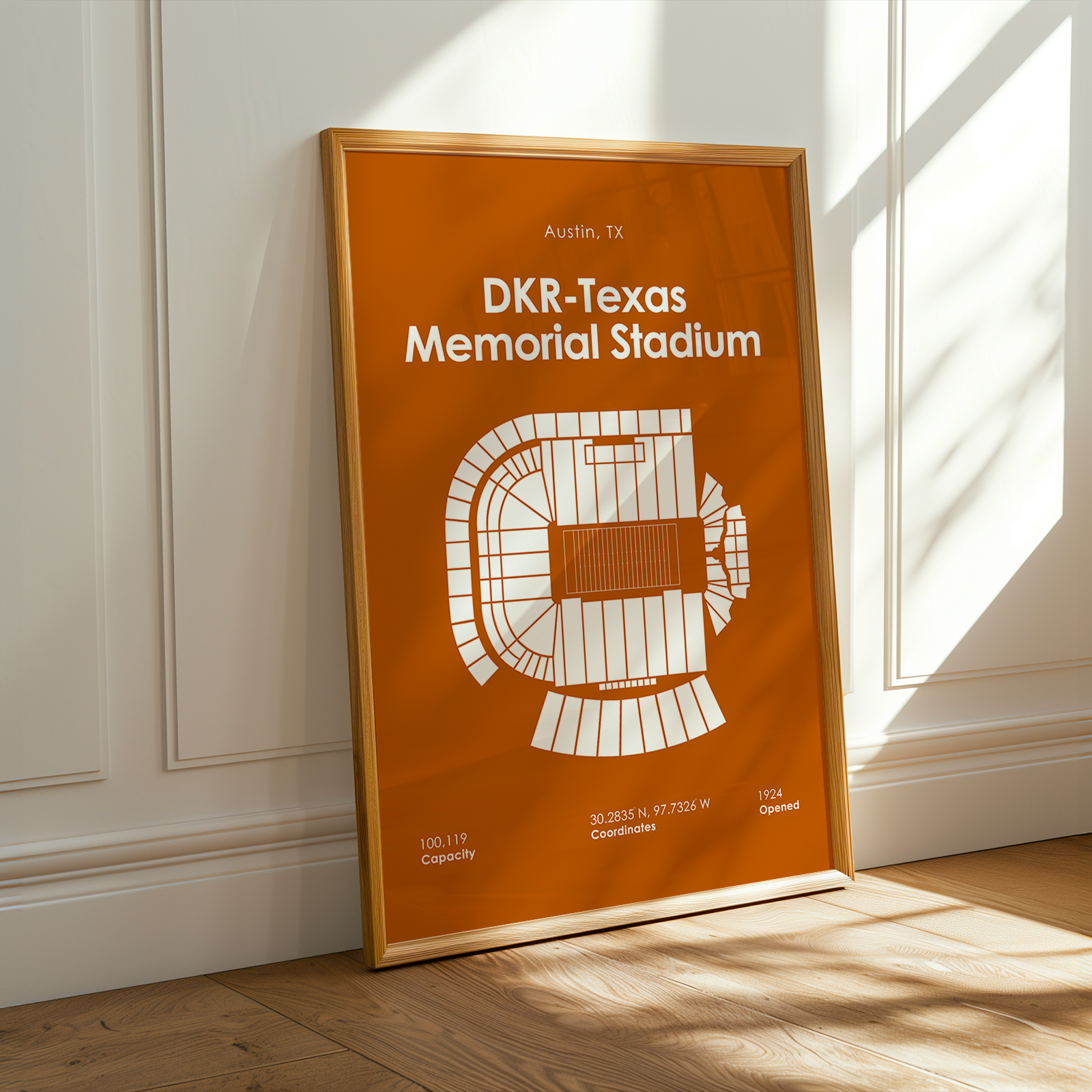 A framed orange and white stadium floor plan print leaning against a white paneled wall on a light wood floor, with warm sunlight and shadows across the scene.