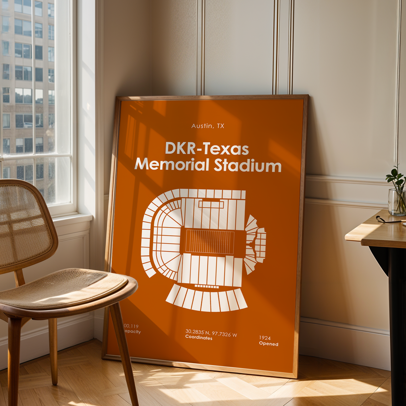 A framed orange and white poster of DKR-Texas Memorial Stadium in Austin, TX, leaning against a wall in a modern room with a wooden chair and desk.