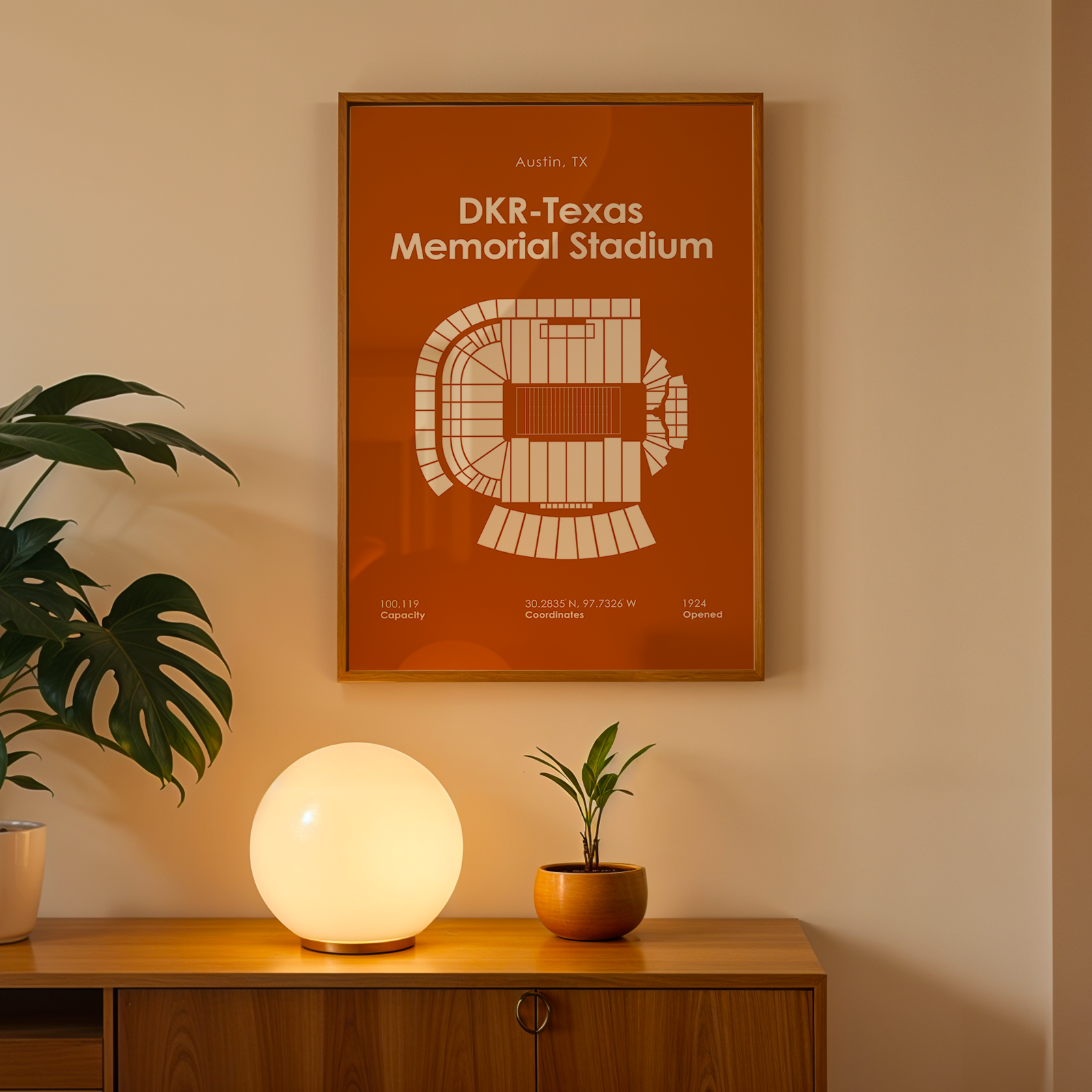 A framed orange and white poster of DKR-Texas Memorial Stadium in Austin, TX, hanging on a beige wall above a wooden console table with a glowing globe lamp and potted plants.