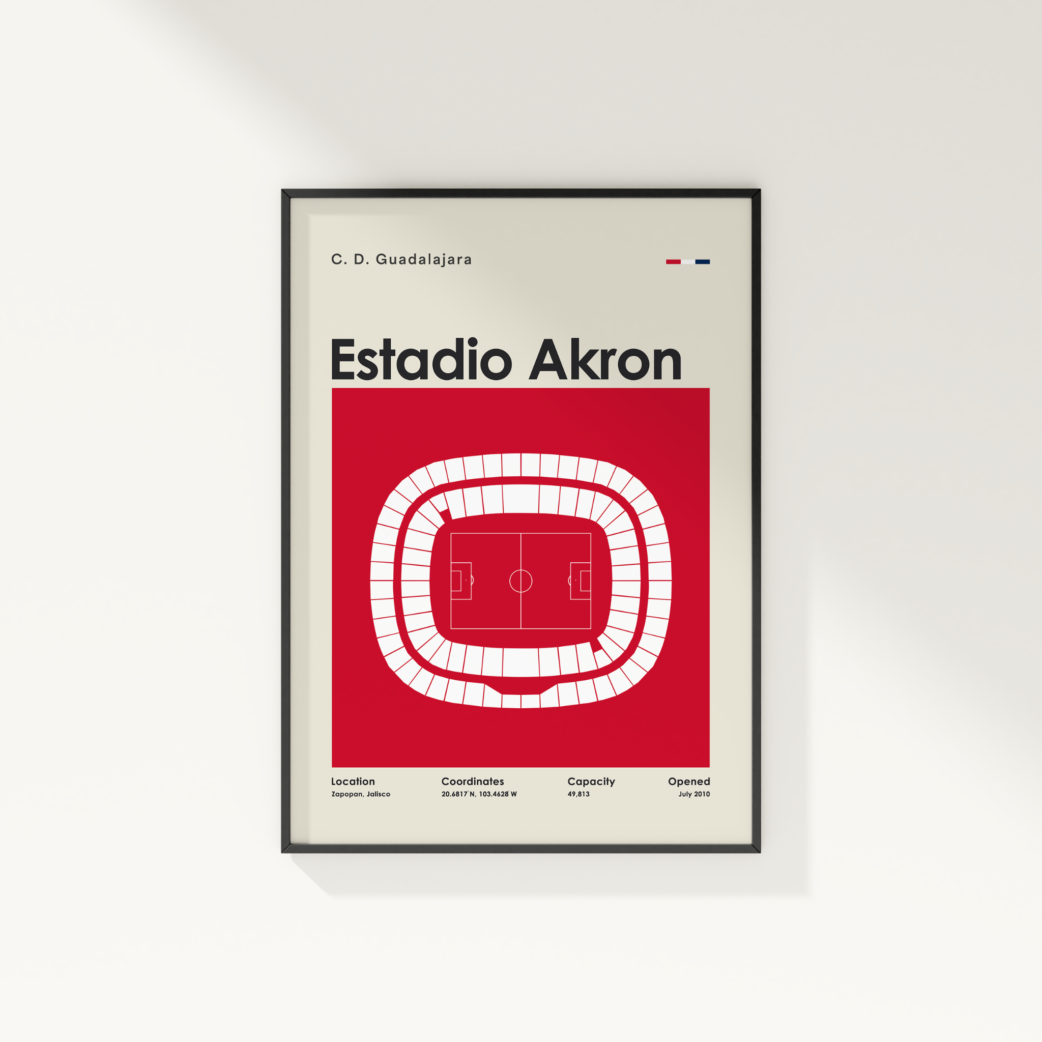 A framed minimalist art print of the Estadio Akron football stadium in Mexico, featuring a red and white stadium layout on a light beige background with a thin black frame, shown hanging on a white wall.