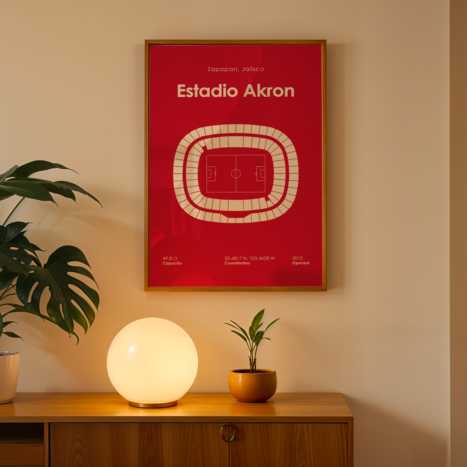 A framed red poster with a white stadium diagram and the text 'Estadio Akron' hangs on a beige wall above a wooden console table. A glowing spherical lamp, a small potted plant, and a larger leafy houseplant sit on the table.