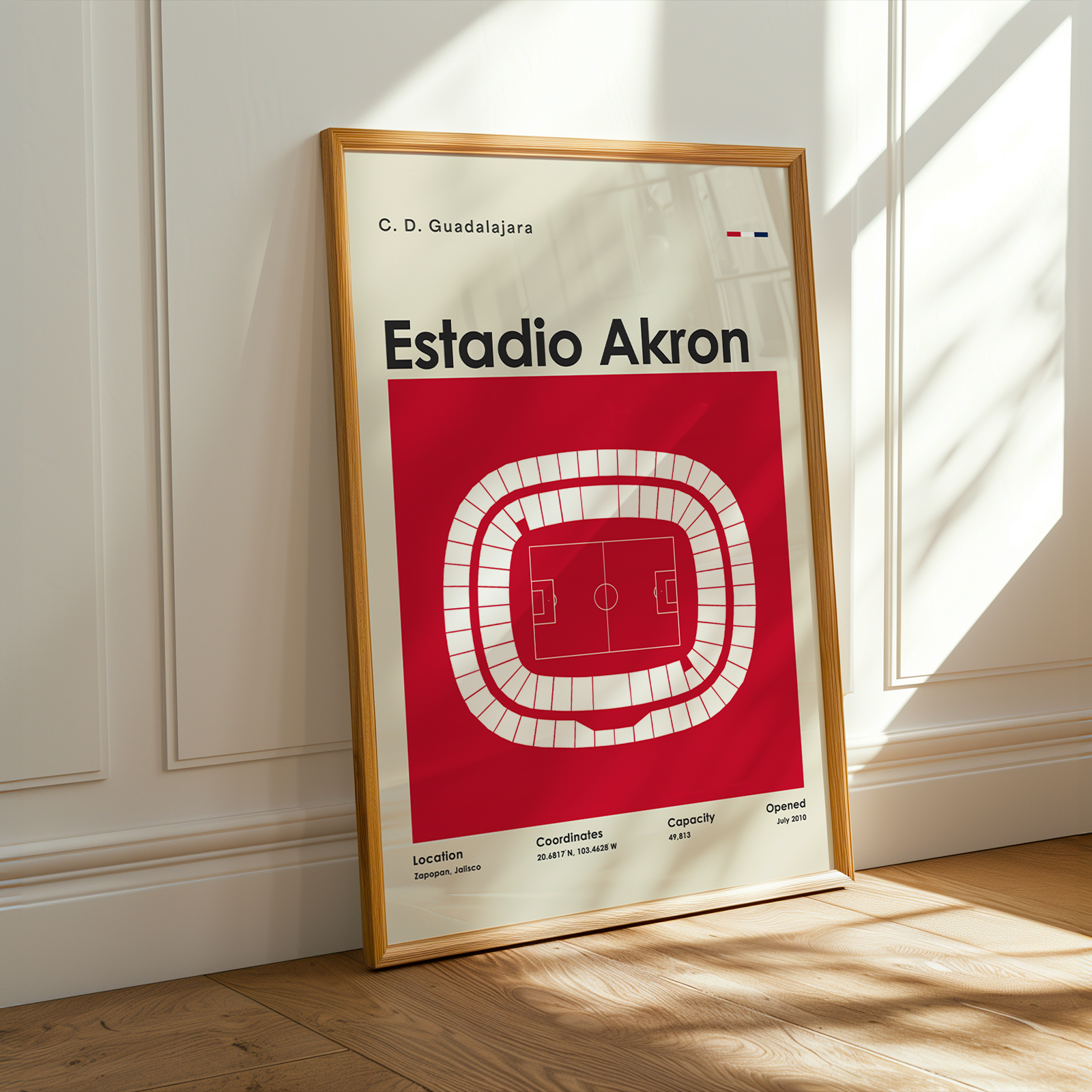 A framed football stadium poster for Estadio Akron leaning against a white paneled wall on a wooden floor, with warm sunlight casting shadows across the scene.