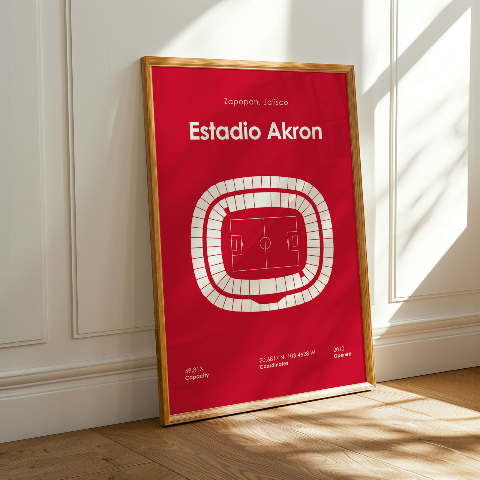 A framed sports poster of Estadio Akron leaning against a white paneled wall on a wooden floor, with warm sunlight casting shadows on the wall and floor.