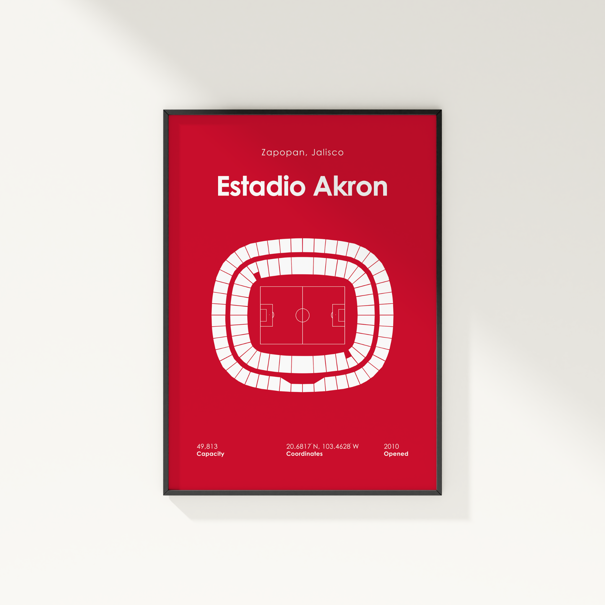 A framed minimalist stadium floor plan poster for Estadio Akron, featuring a red background with white text and a white stadium diagram, mounted on a light gray wall.