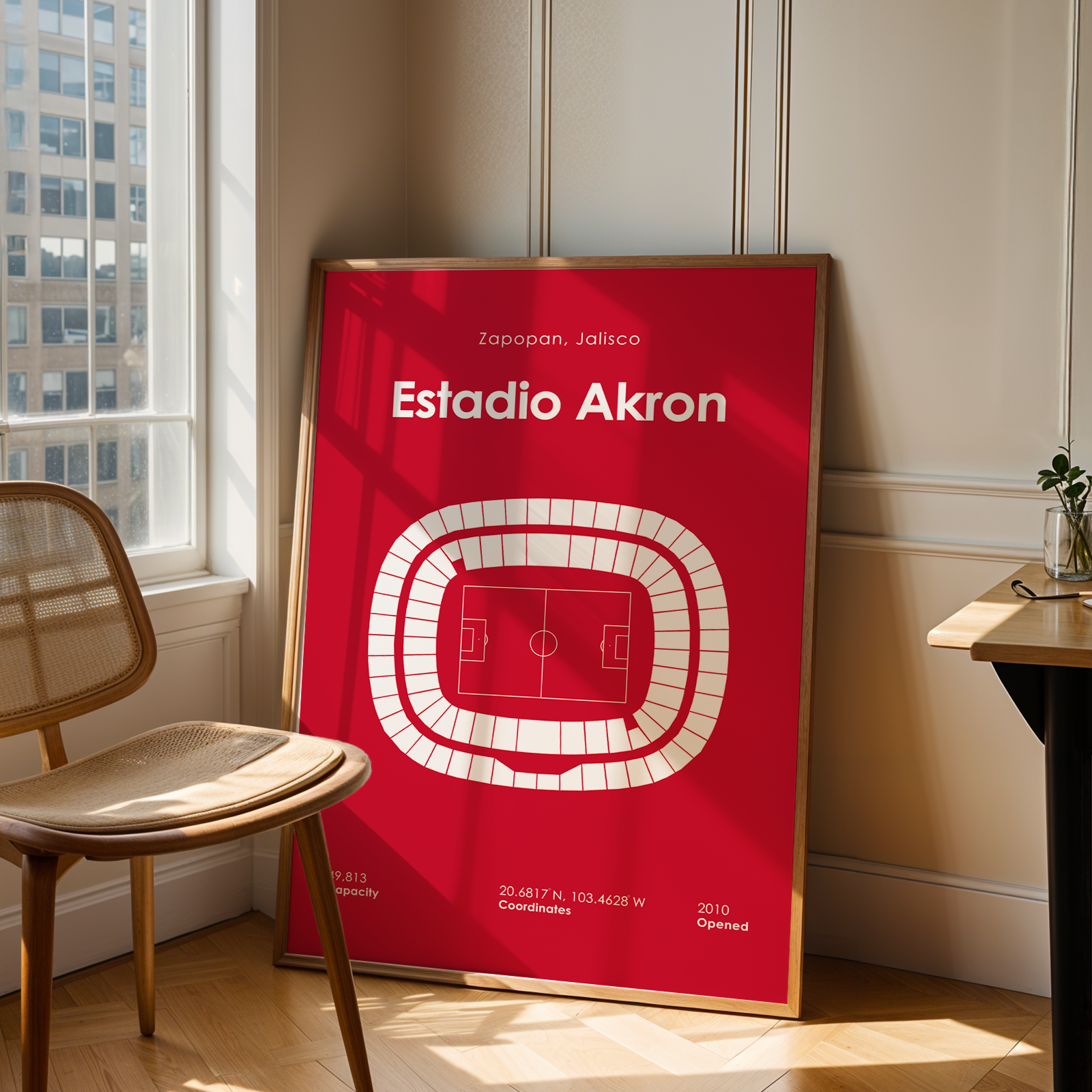 A framed red sports poster leaning against a wall in a modern room, featuring a white stadium layout and the text 'Estadio Akron'. A wooden chair is on the left and a desk with a plant is on the right.
