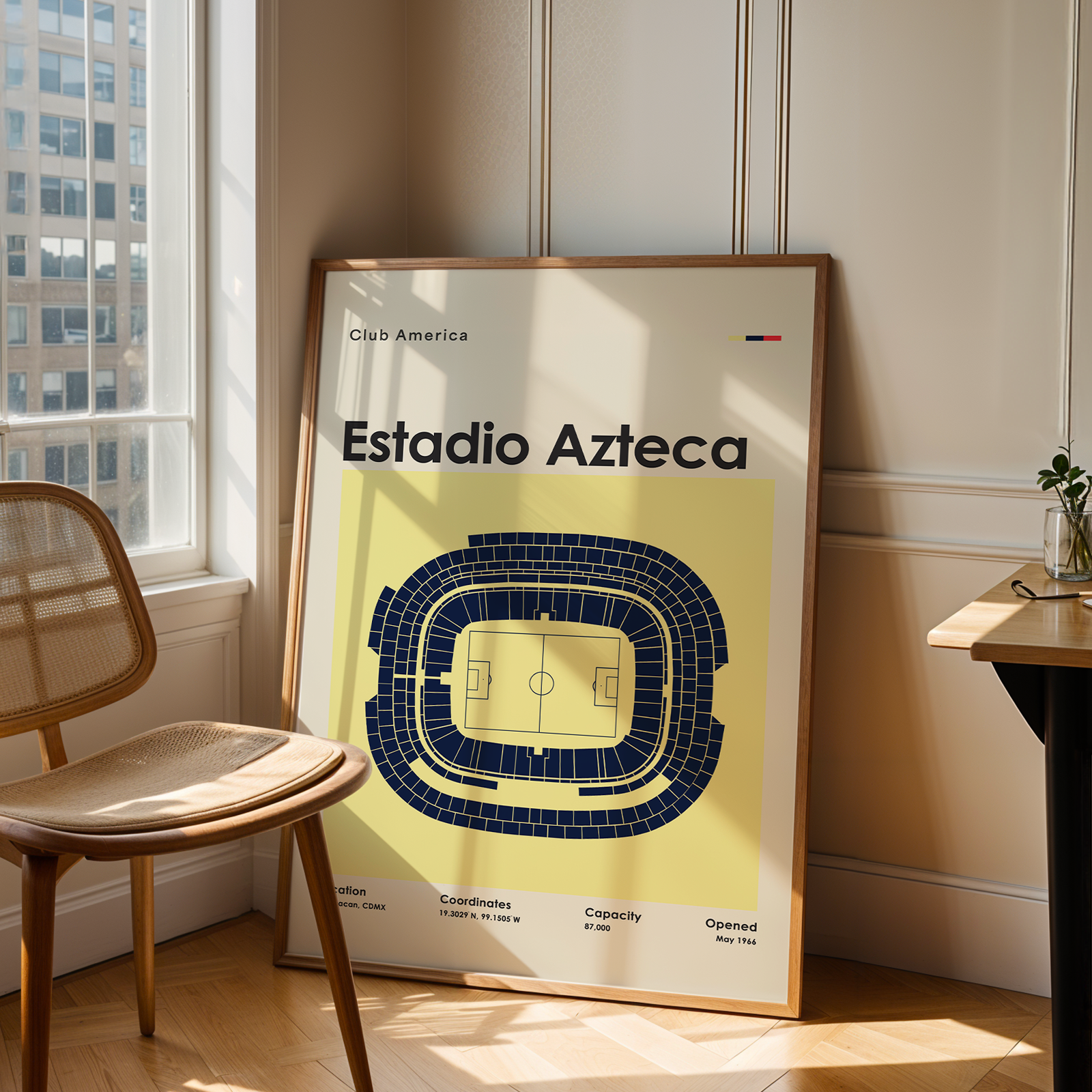 A framed poster leaning against a wall in a sunlit room, showing a stadium floor plan labeled 'Estadio Azteca' with 'Club America' text. A wooden chair is beside the window and a small desk sits on the right.