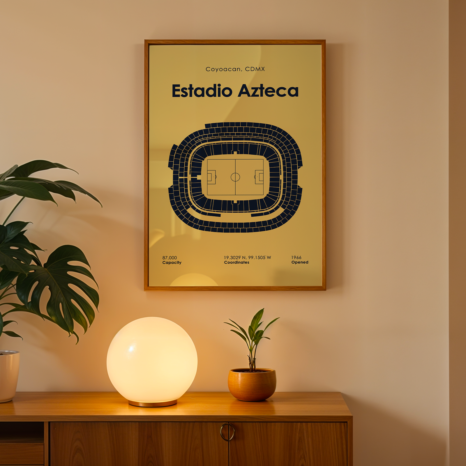 A framed Estadio Azteca sports seating chart poster hangs on a beige wall above a wooden console table. The print features a dark blue stadium diagram on a warm cream background, enclosed in a light wood frame.