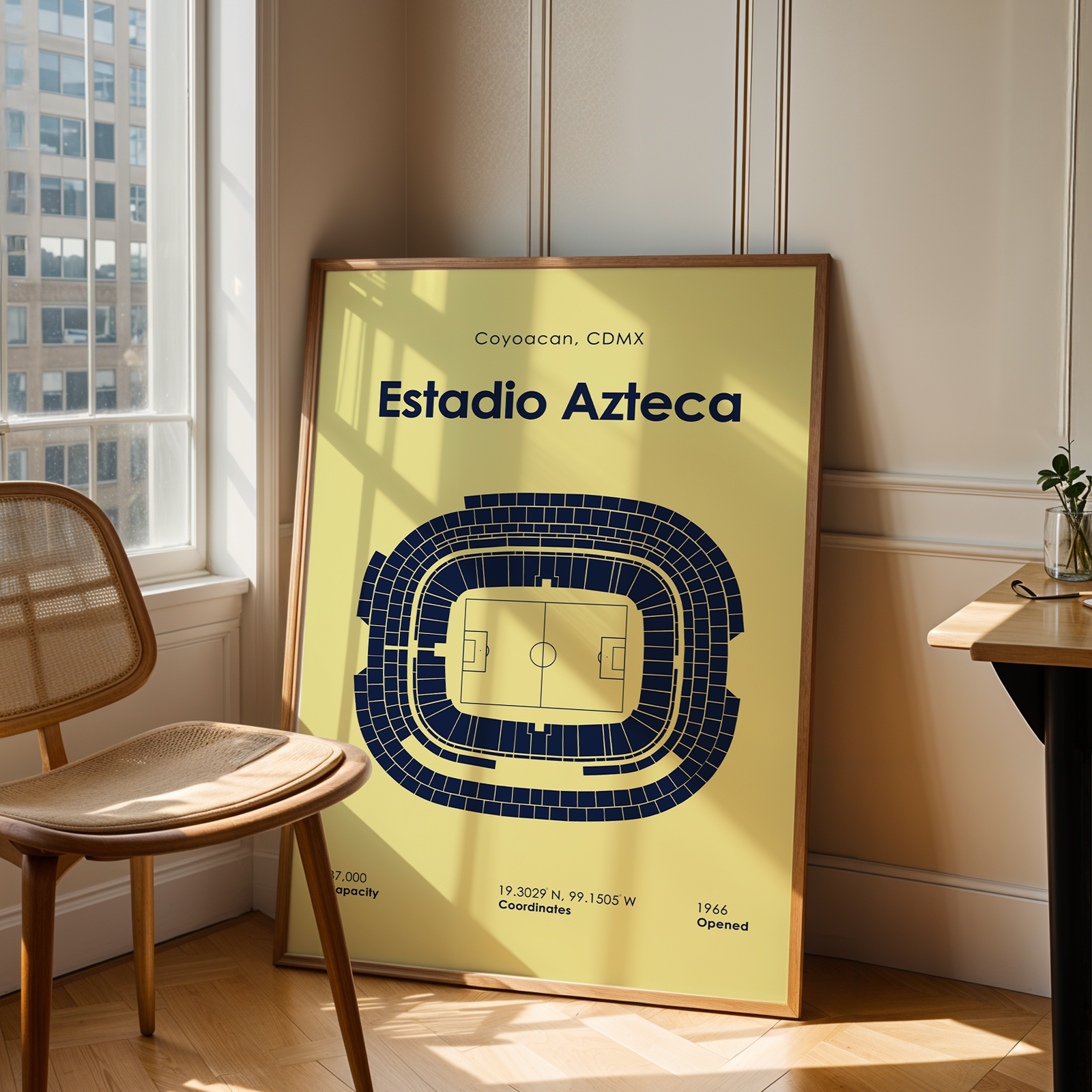 A framed art print of the Estadio Azteca stadium in Mexico, featuring a yellow background and a dark blue stadium diagram, leaning against a beige wall in a modern room next to a wooden chair and desk.