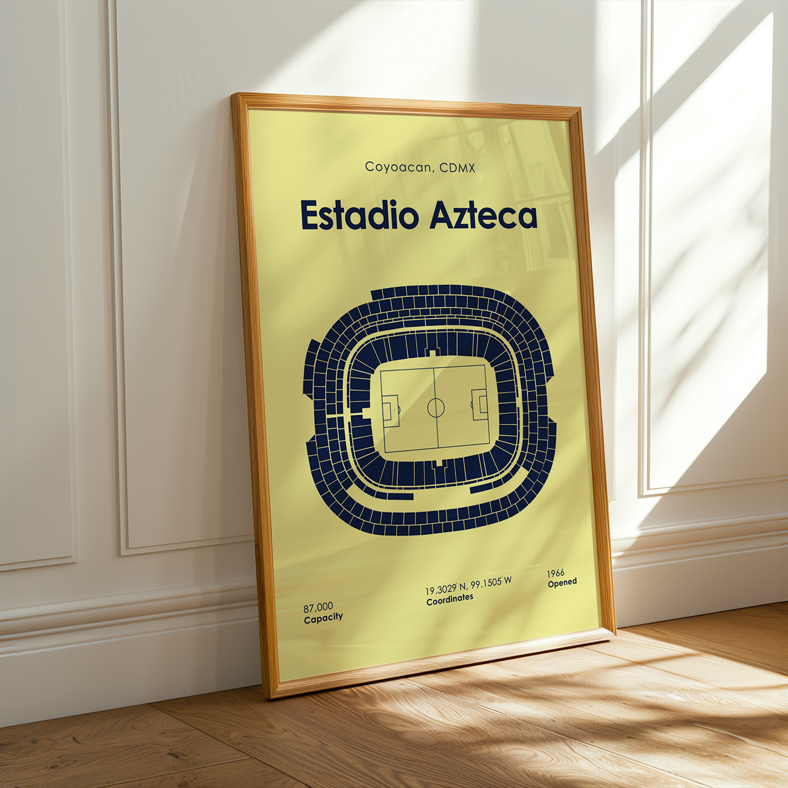 A framed stadium floor plan poster for Estadio Azteca in Coyoacán, CDMX, leaning against a white paneled wall on a wooden floor with warm sunlight streaming in from a window.