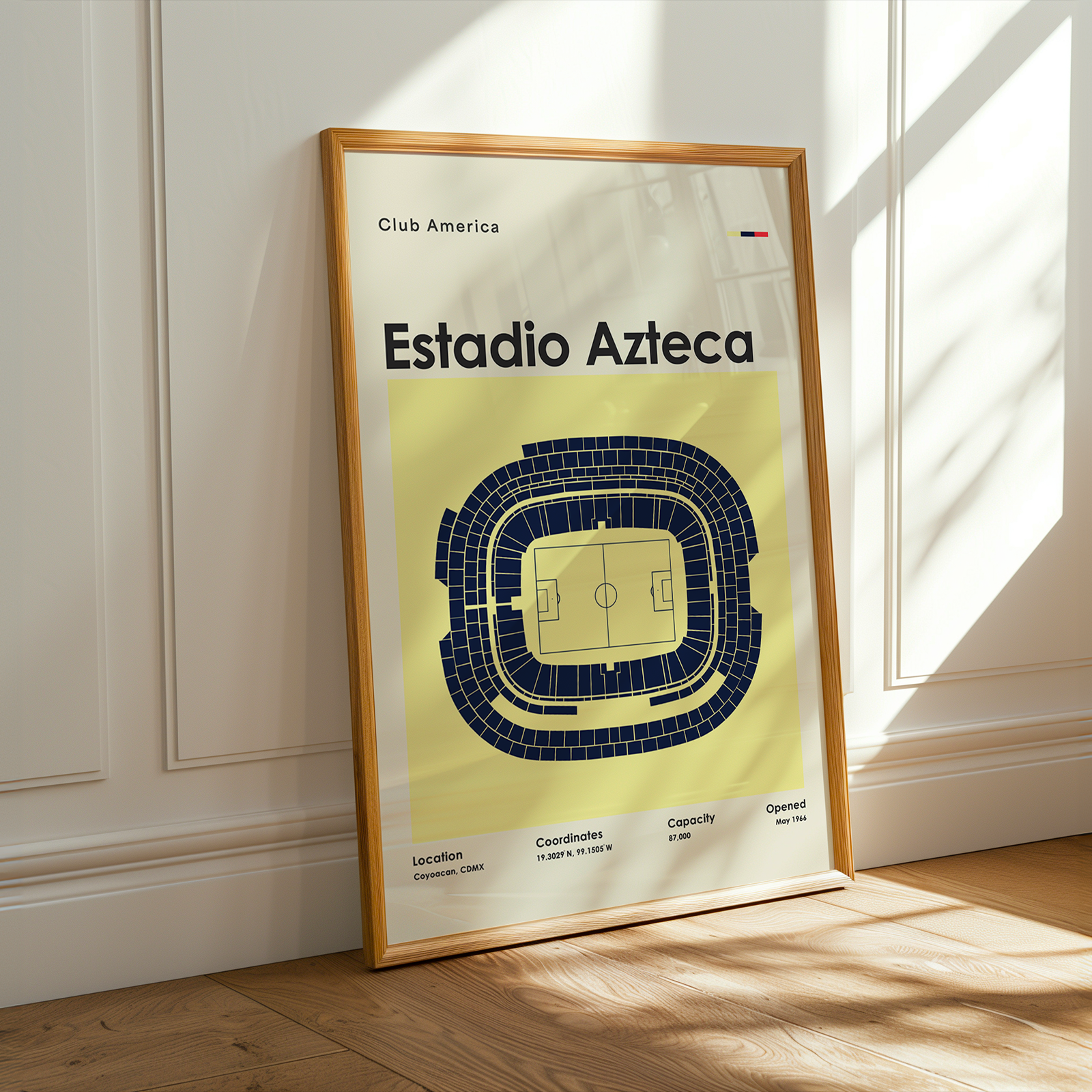 A framed poster of the Estadio Azteca stadium layout of Club America, leaning against a white paneled wall on a wooden floor with warm sunlight streaming in.