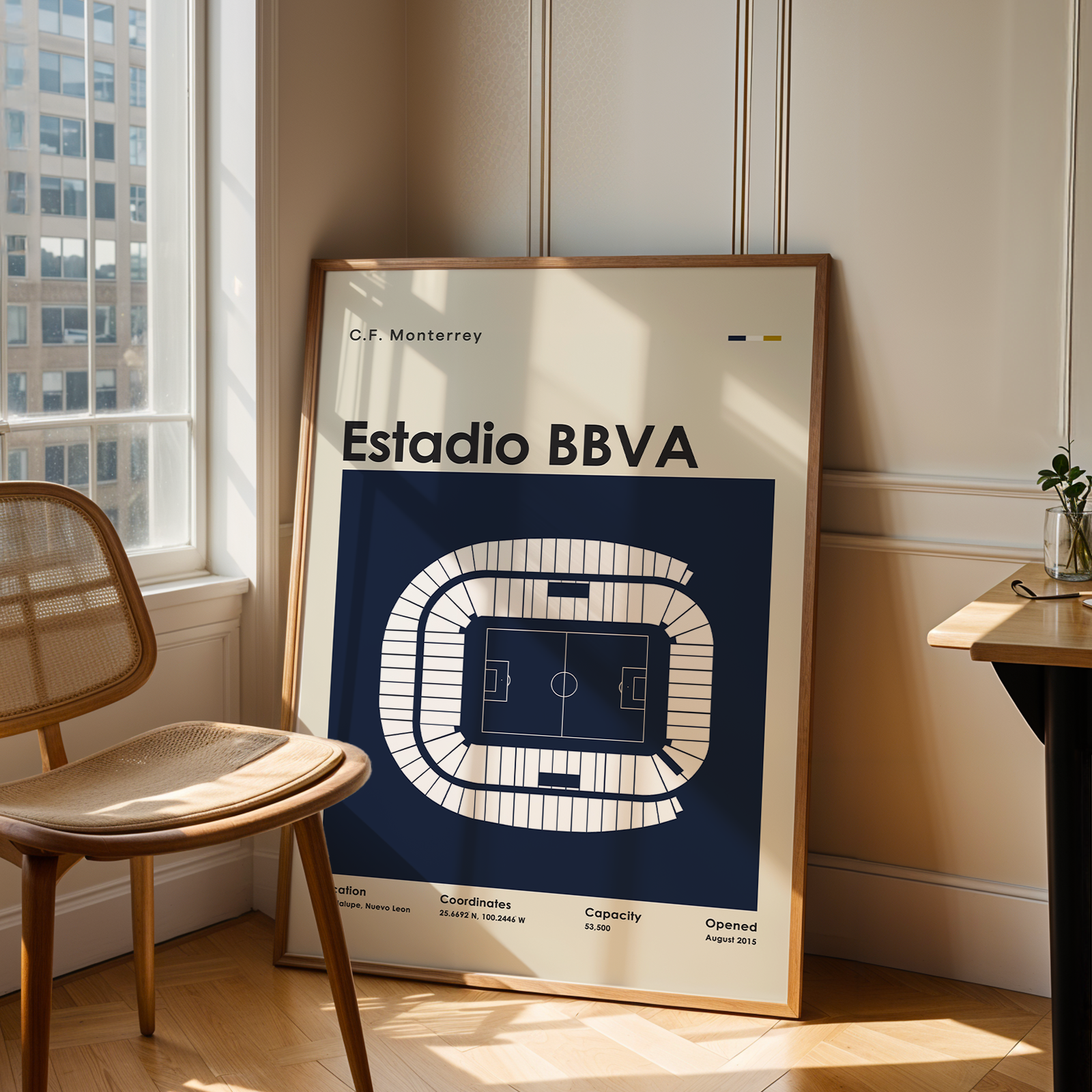 A framed art print of the Estadio BBVA stadium layout, leaning against a beige wall in a modern room next to a wooden chair and desk.