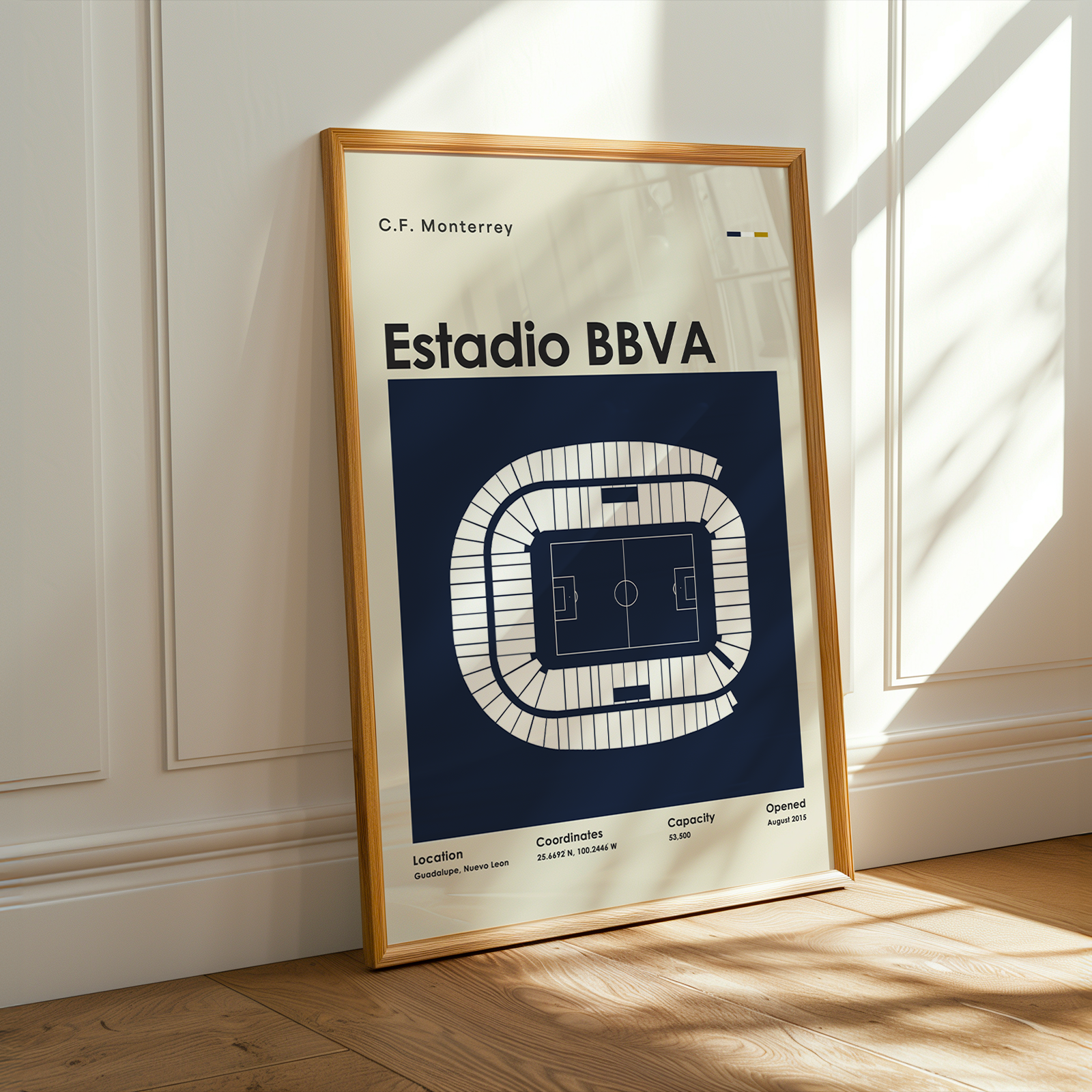 A framed poster of the Estadio BBVA football stadium in Monterrey, featuring a top-down field diagram on a navy blue background. The poster is framed in light wood and leans against a white paneled wall on a wooden floor, with warm sunlight casting shadows across the scene.