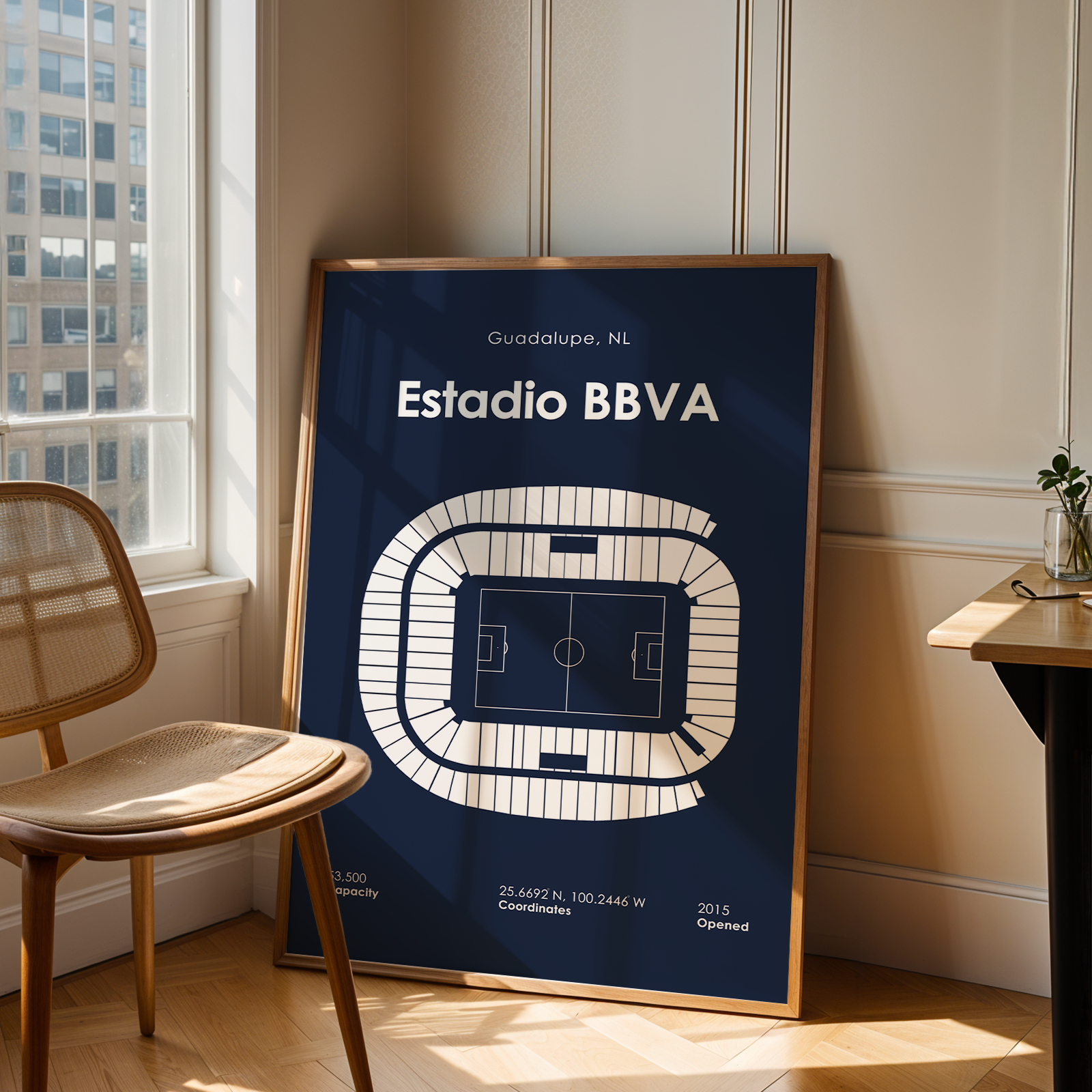 A framed vertical poster of the Estadio BBVA football stadium in Guadalupe, NL, leaning against a beige wall in a modern room next to a wooden chair and desk.