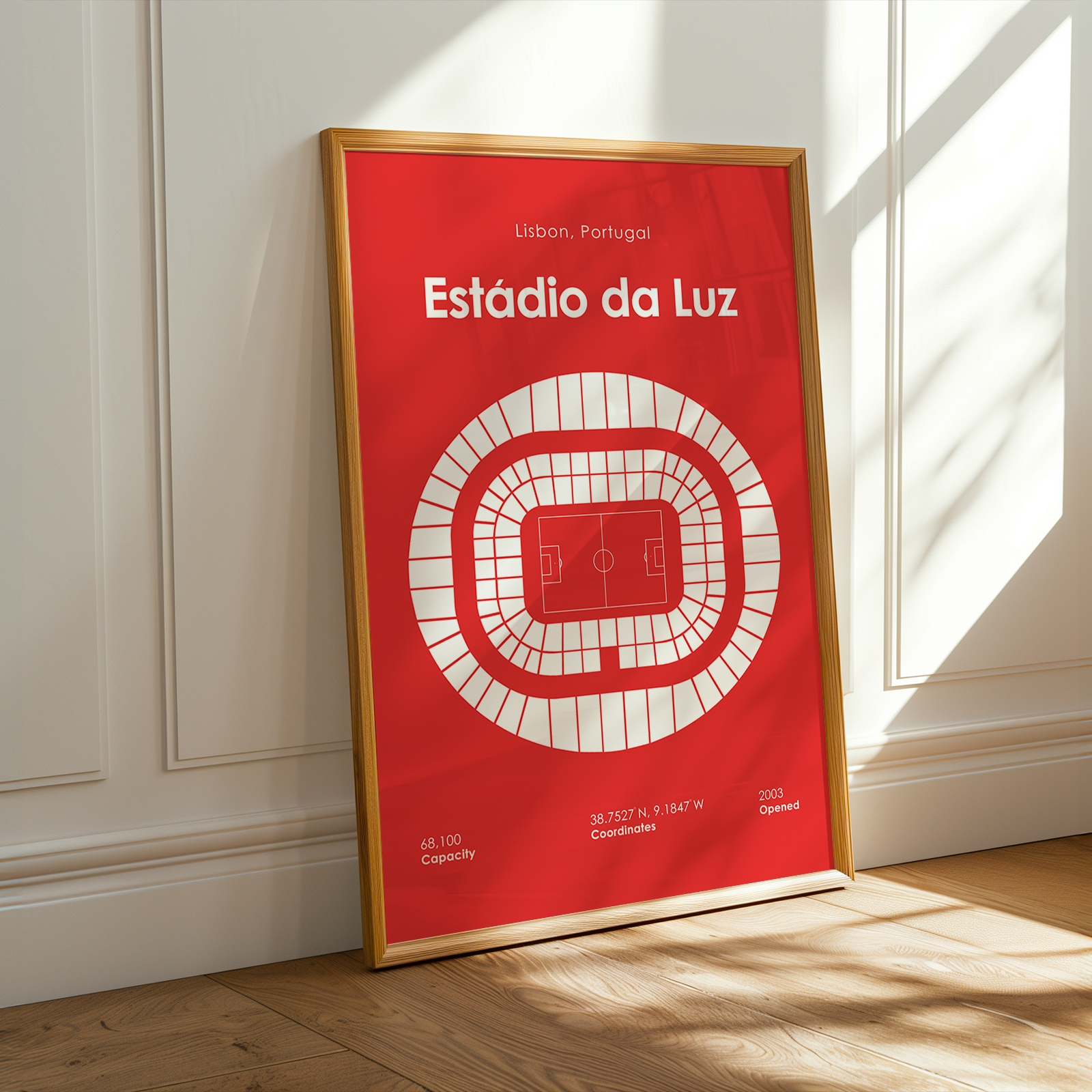 A framed minimalist stadium map poster of Estãdio da Luz in Lisbon, Portugal, leaning against a white paneled wall on a light wood floor with warm sunlight streaming in.
