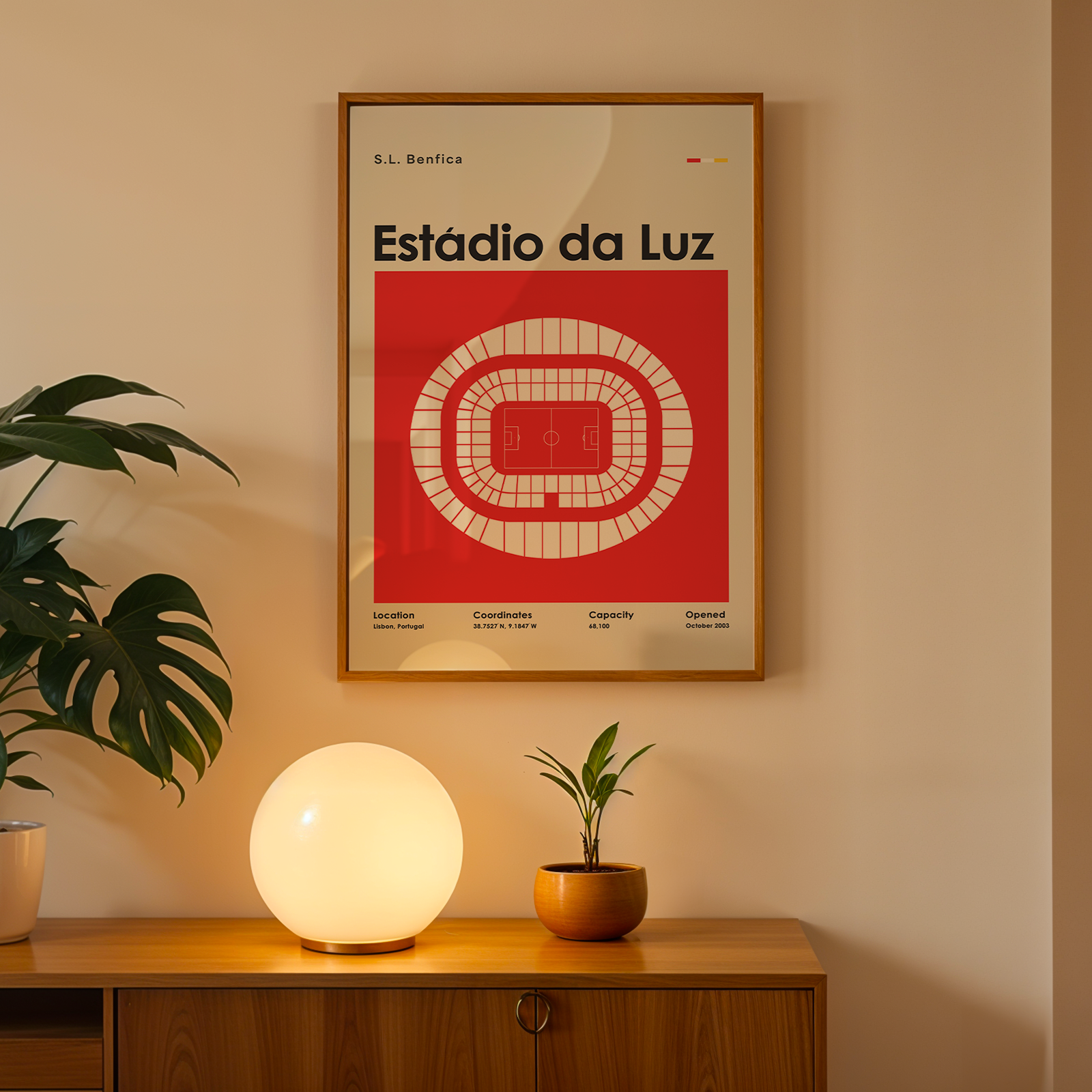 A framed vintage-style poster of the Estãdio da Luz stadium in Lisbon, featuring a bold red and cream graphic design, hanging on a beige wall above a wooden console table with a glowing globe lamp and potted plants.