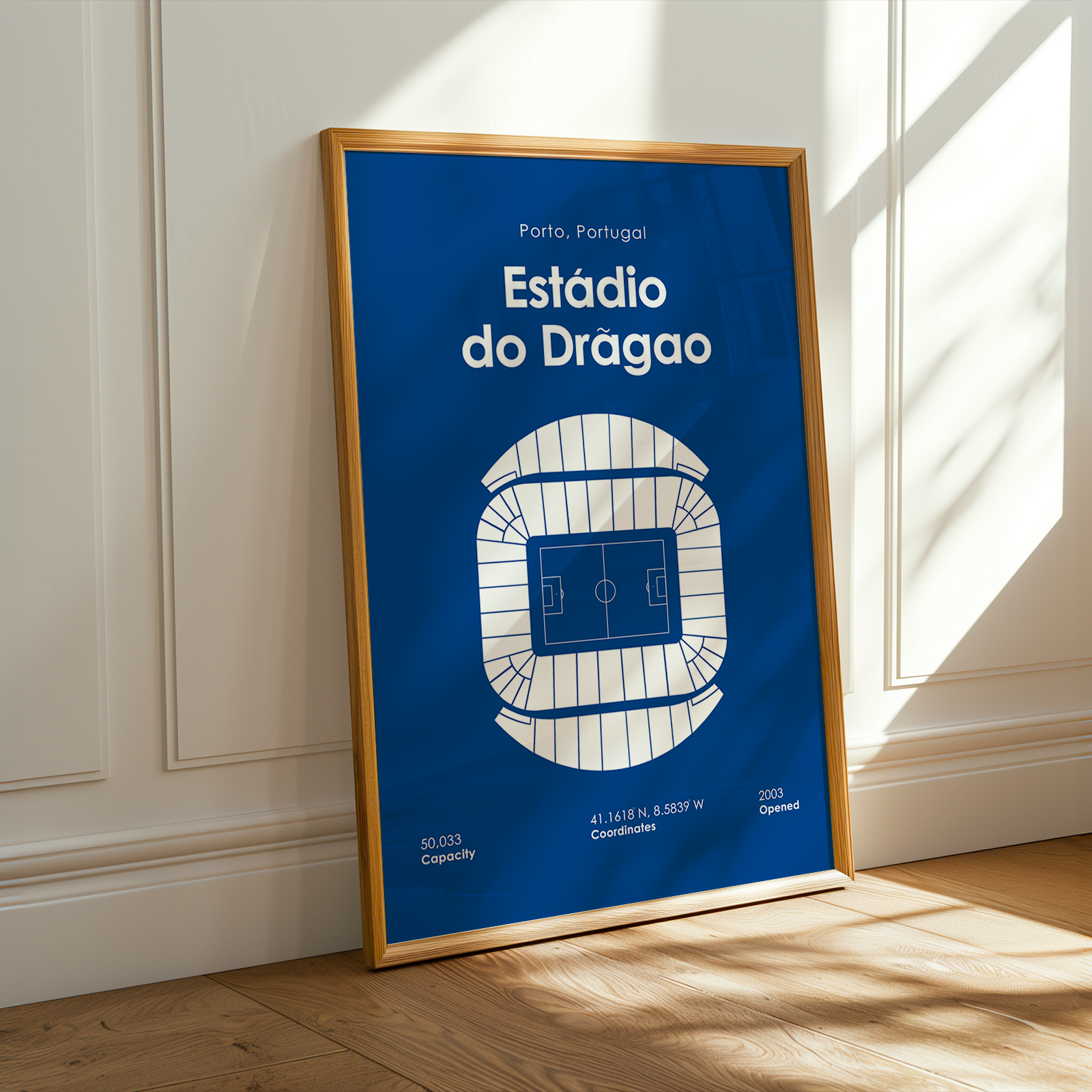 A framed vertical art print of the Estãdio do Dragao stadium in Portugal, featuring a blue and white schematic diagram of the stadium layout. The print is housed in a light wood frame and leans against a white paneled wall on a light wood floor, with warm sunlight casting shadows on the wall and floor.