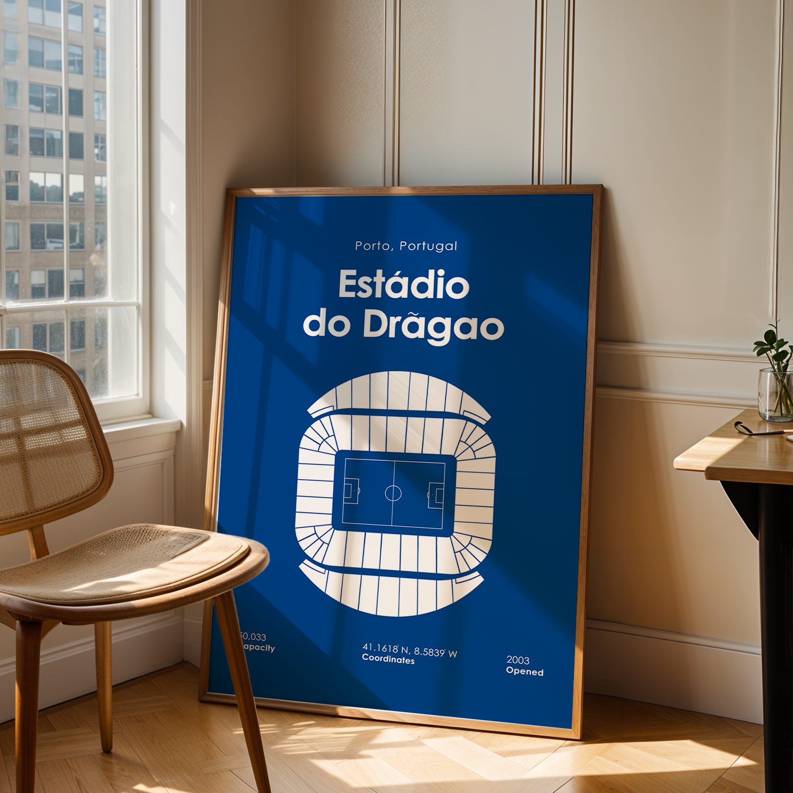 A framed vertical poster leaning against a wall in a sunlit room. The poster features a blue background with white text reading 'Porto, Portugal' and 'Estádio do Dragao,' plus a white stadium outline graphic.