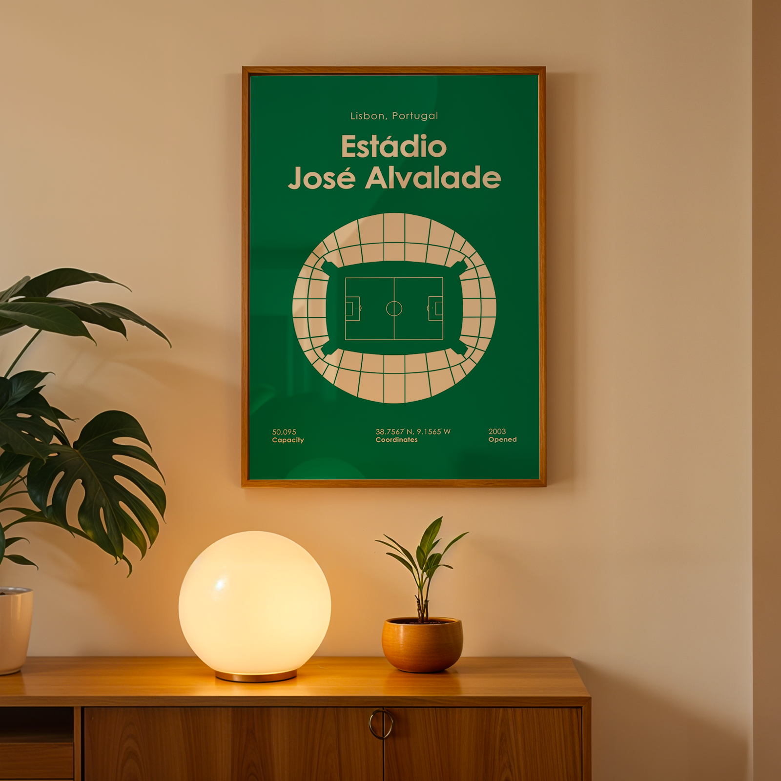 A framed green poster with white text reading 'Estadio José Alvalade' hangs on a beige wall above a wooden console table. A glowing spherical lamp, a small potted plant, and a larger leafy houseplant sit on the table in a warm, styled interior.