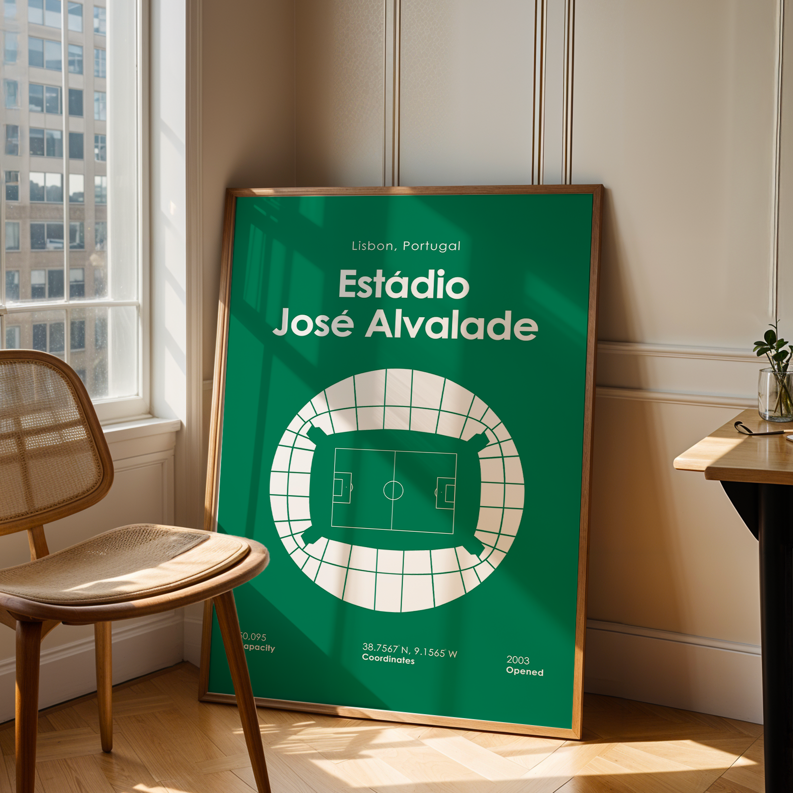A framed green sports poster leaning against a wall in a sunlit room, next to a wooden chair and a small table with a plant. The poster reads "Estãdio José Alvalade" and shows a stadium floor plan.
