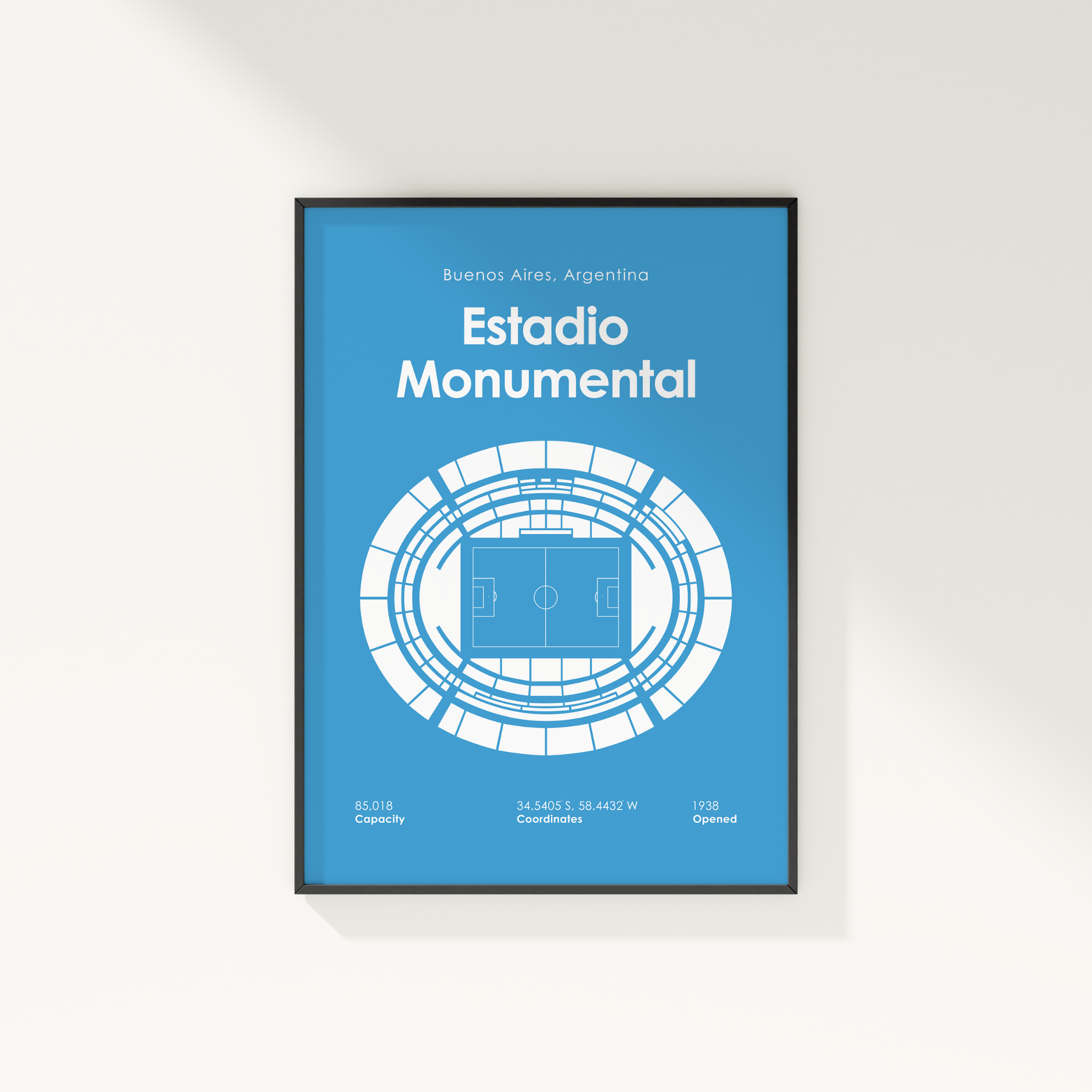 A framed poster print featuring a blue and white schematic design of Estadio Monumental in Buenos Aires, Argentina, displayed on a light beige wall.