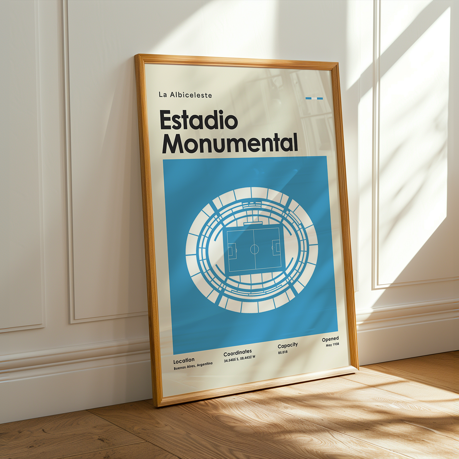 A framed poster leaning against a white paneled wall on a wooden floor, with warm sunlight and window shadows cast across the scene. The poster reads 'La Albiceleste Estadio Monumental' with a blue stadium graphic and a light wood frame.