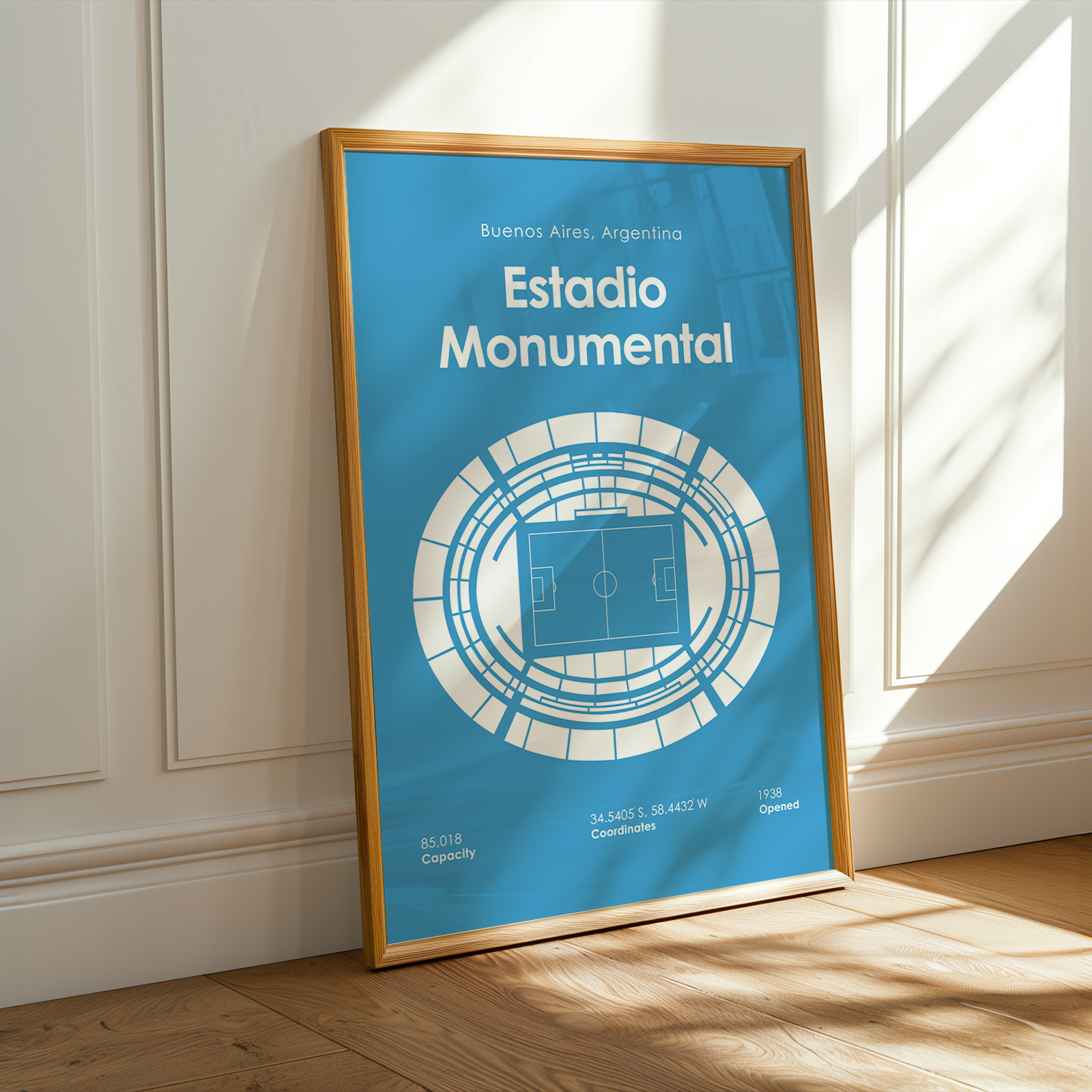 A framed art print of the Estadio Monumental in Buenos Aires, Argentina, leaning against a white paneled wall on a wooden floor with warm sunlight and window shadows.