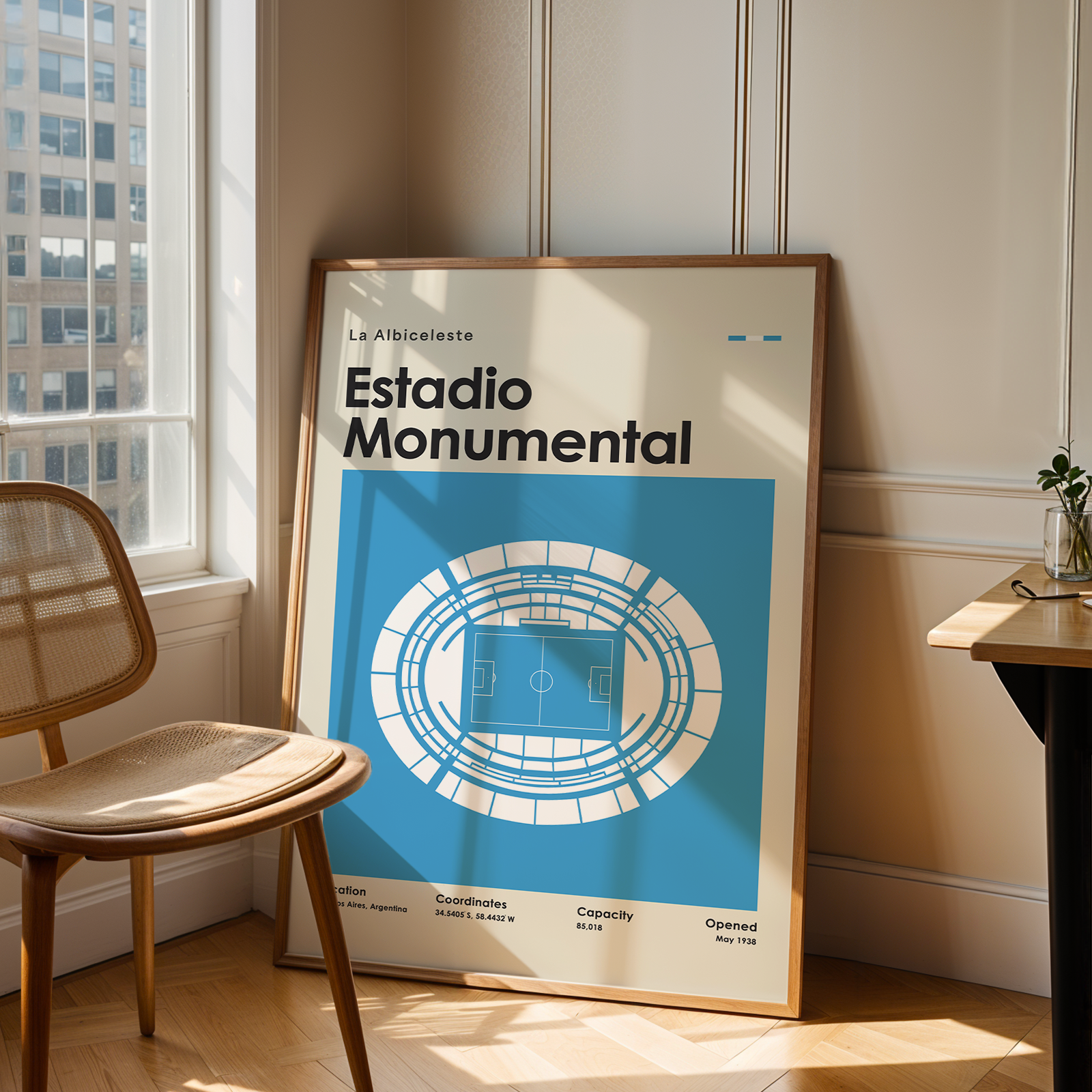 A framed poster leaning against a wall in a sunlit room, showing a blue and white stadium illustration with the text 'Estadio Monumental.' A wooden chair sits to the left, and a small table with a plant is to the right.