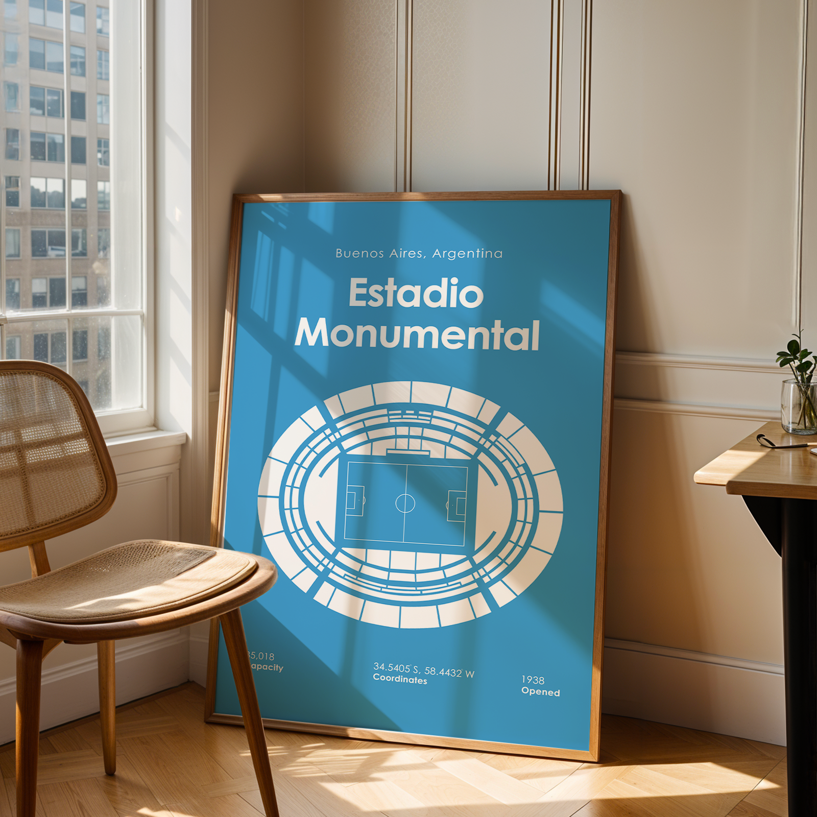 A framed poster leaning against a wall in a sunlit room, showing a blue stadium floor plan with the text 'Estadio Monumental' and 'Buenos Aires, Argentina.' A wooden chair is beside the window, and a small table sits on the right.