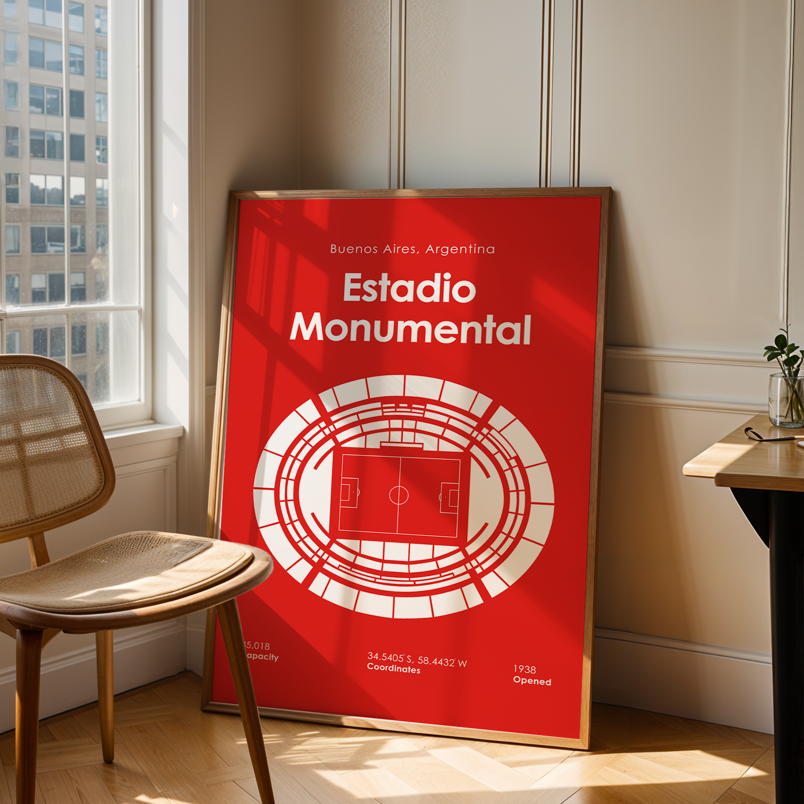 A framed red and white poster of Estadio Monumental in Buenos Aires, Argentina, leaning against a beige paneled wall in a sunlit room next to a wooden chair and desk.