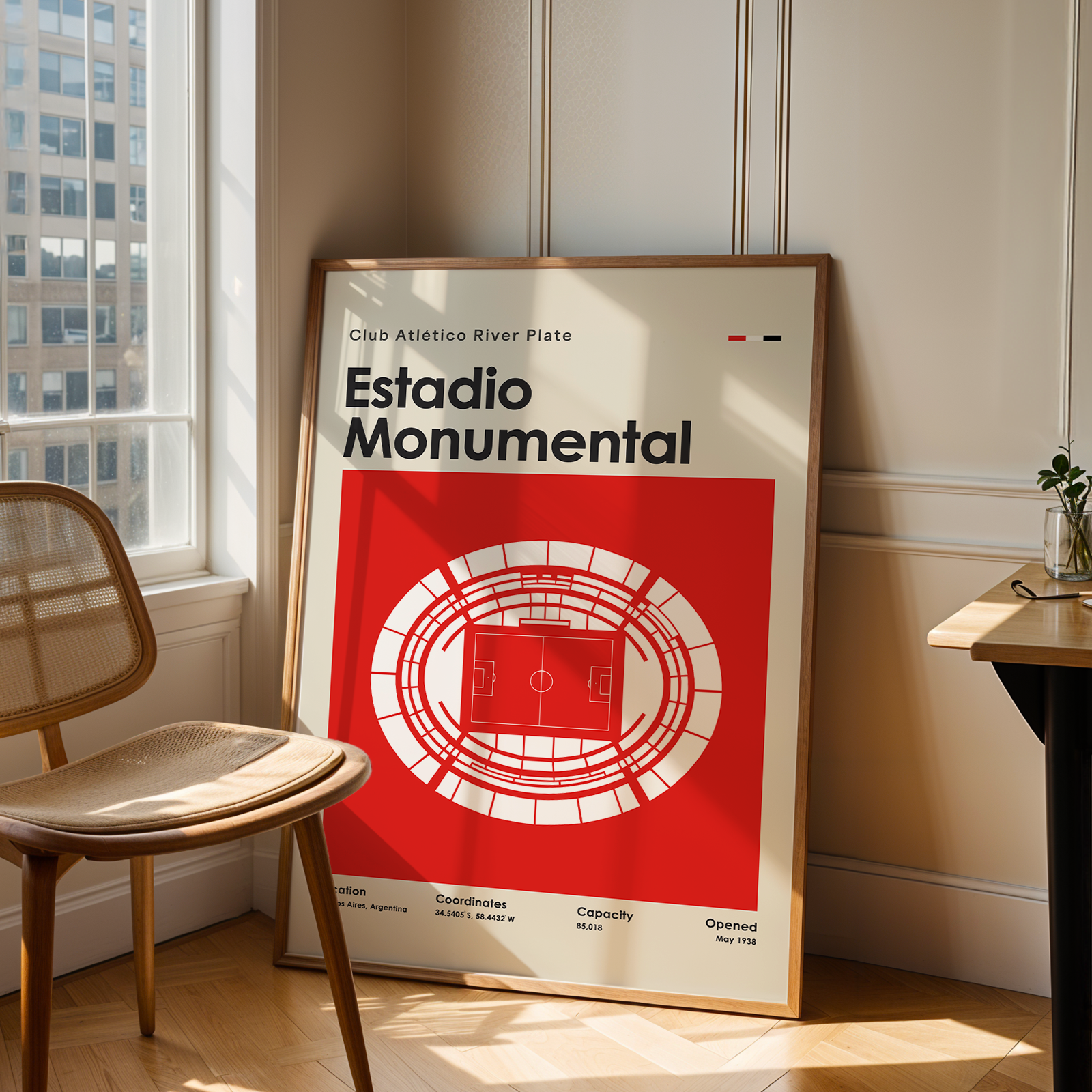 A framed poster of Estadio Monumental in Buenos Aires, featuring a bold red and white stadium design, leaning against a wall in a sunlit room next to a wooden chair and desk.