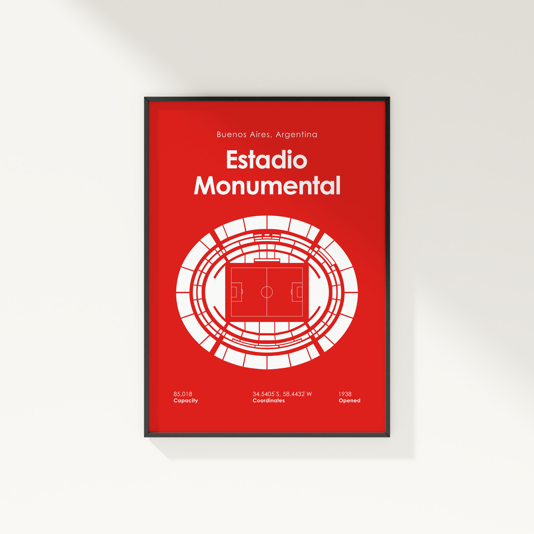 A minimalist red and white poster of the Estadio Monumental in Buenos Aires, Argentina, shown as a framed print on a light gray wall.
