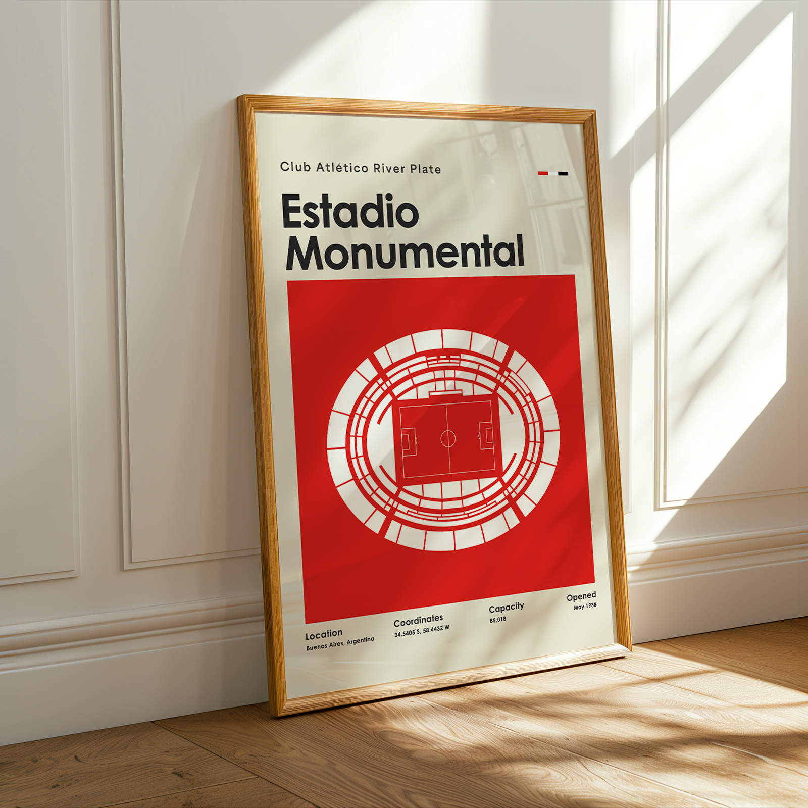 A framed vintage-style football stadium poster leaning against a white paneled wall on a light wood floor, with warm sunlight casting shadows across the scene.