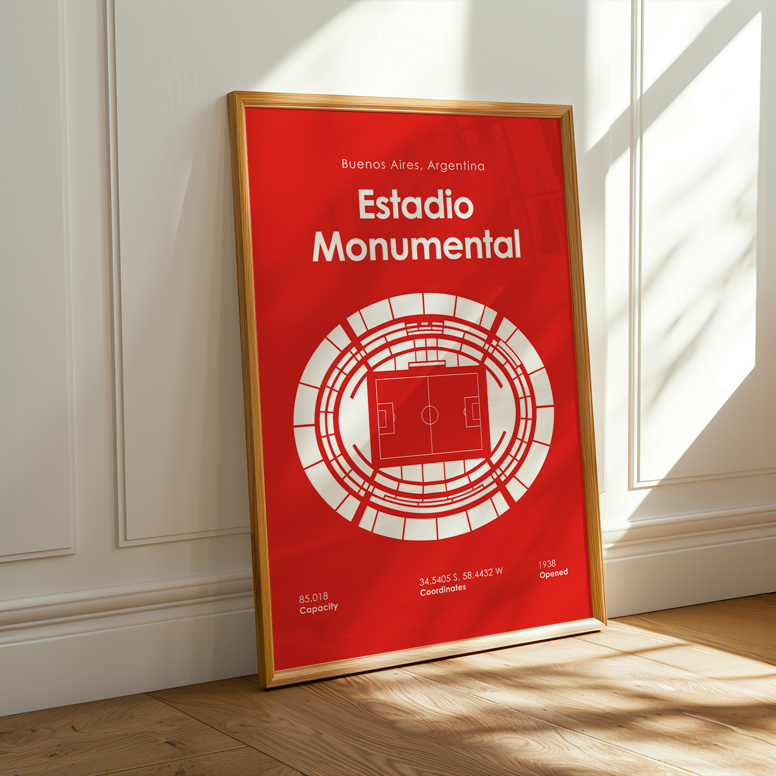 A framed red and white poster of Estadio Monumental in Buenos Aires, Argentina, leaning against a white paneled wall on a light wood floor with warm sunlight and window shadows.