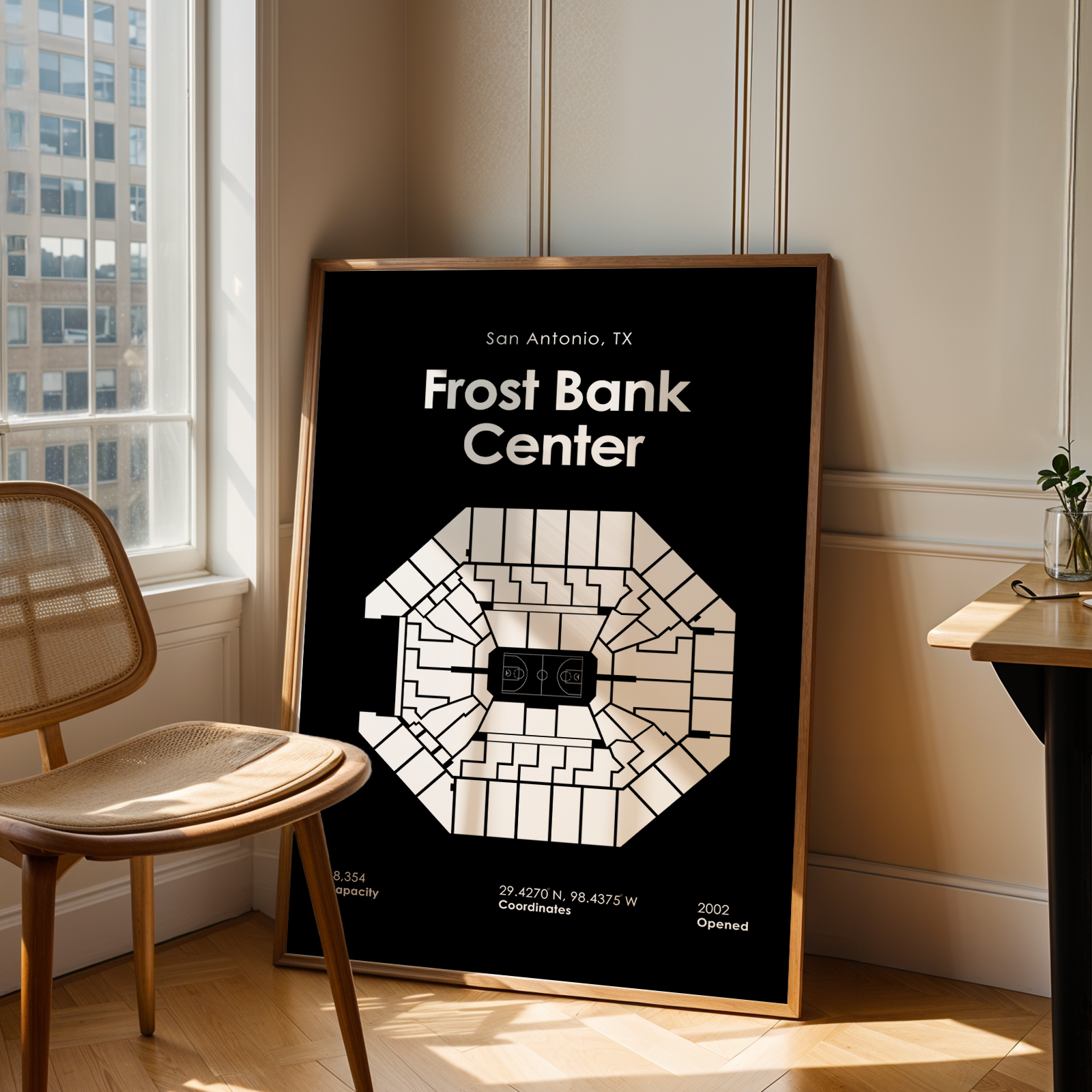 Framed poster of Frost Bank Center in San Antonio, TX, leaning against a wall in a room with a chair and table.