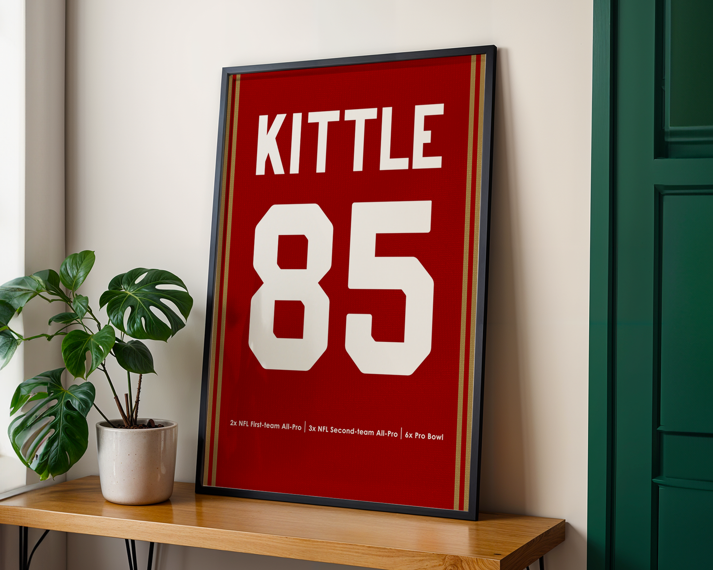 Framed poster with 'Kittle 85' on a wooden shelf next to a plant