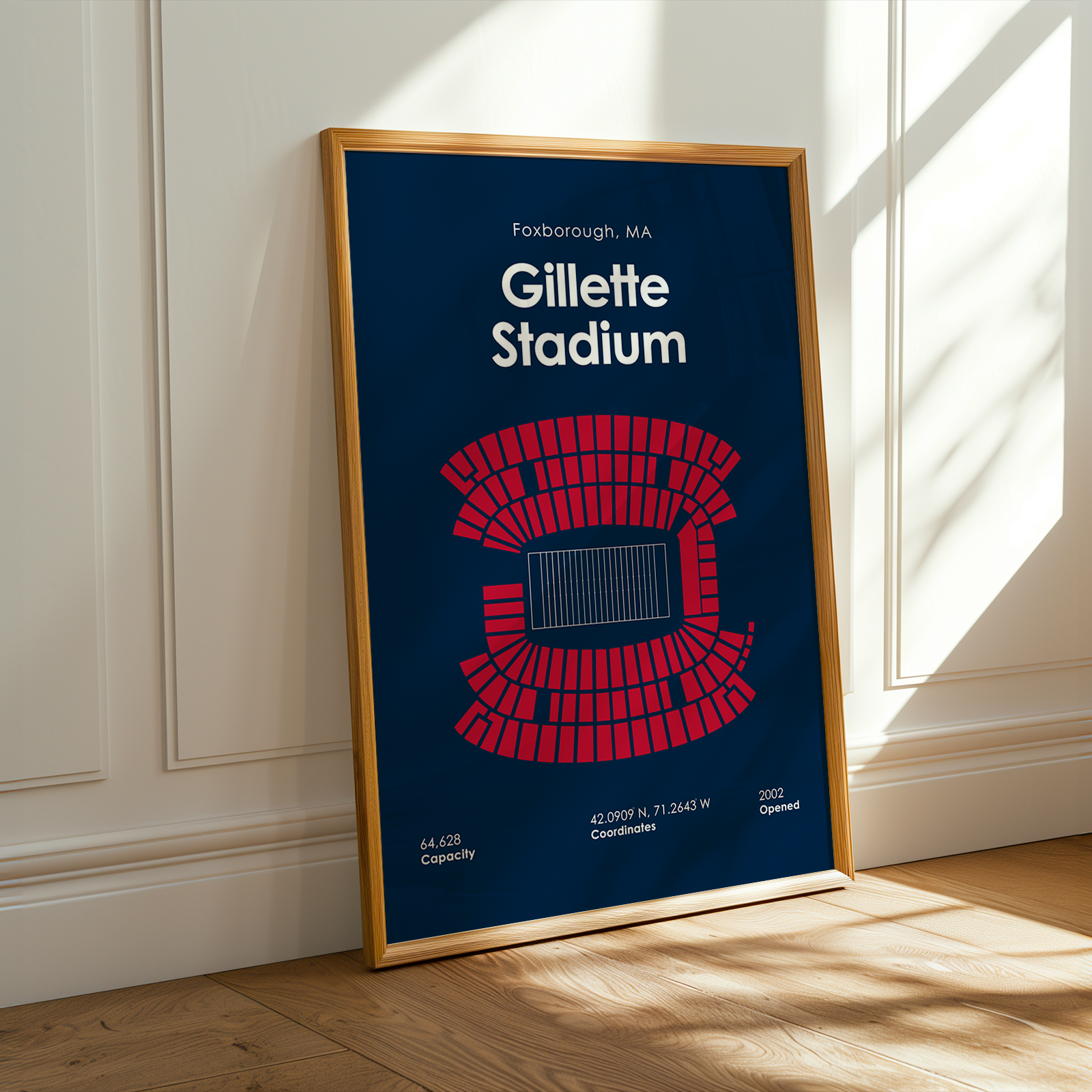 Framed poster of Gillette Stadium with a blue background and red seating diagram, leaning against a white wall.