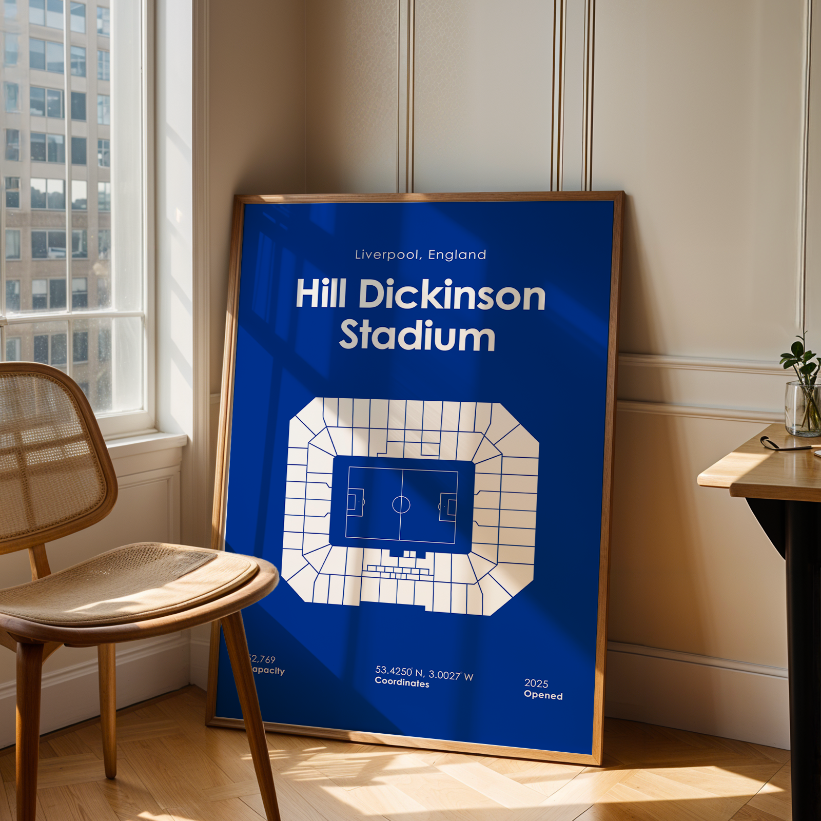 Framed poster of Hill Dickinson Stadium in Liverpool, England, leaning against a wall in a room with a chair and window.
