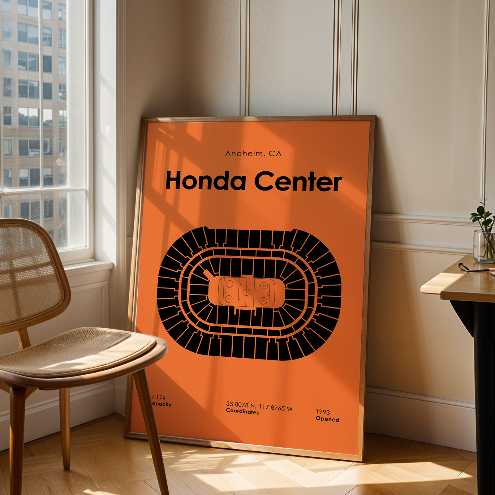 Framed poster of the Honda Center in Anaheim, CA on an orange background in a room with a chair and window.