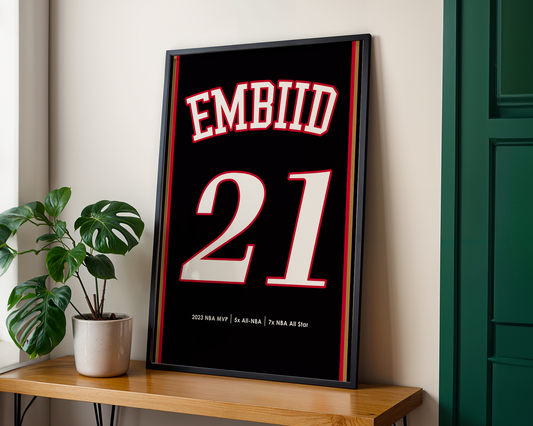 Framed sports jersey with 'EMBIID 21' on a wooden shelf next to a plant.