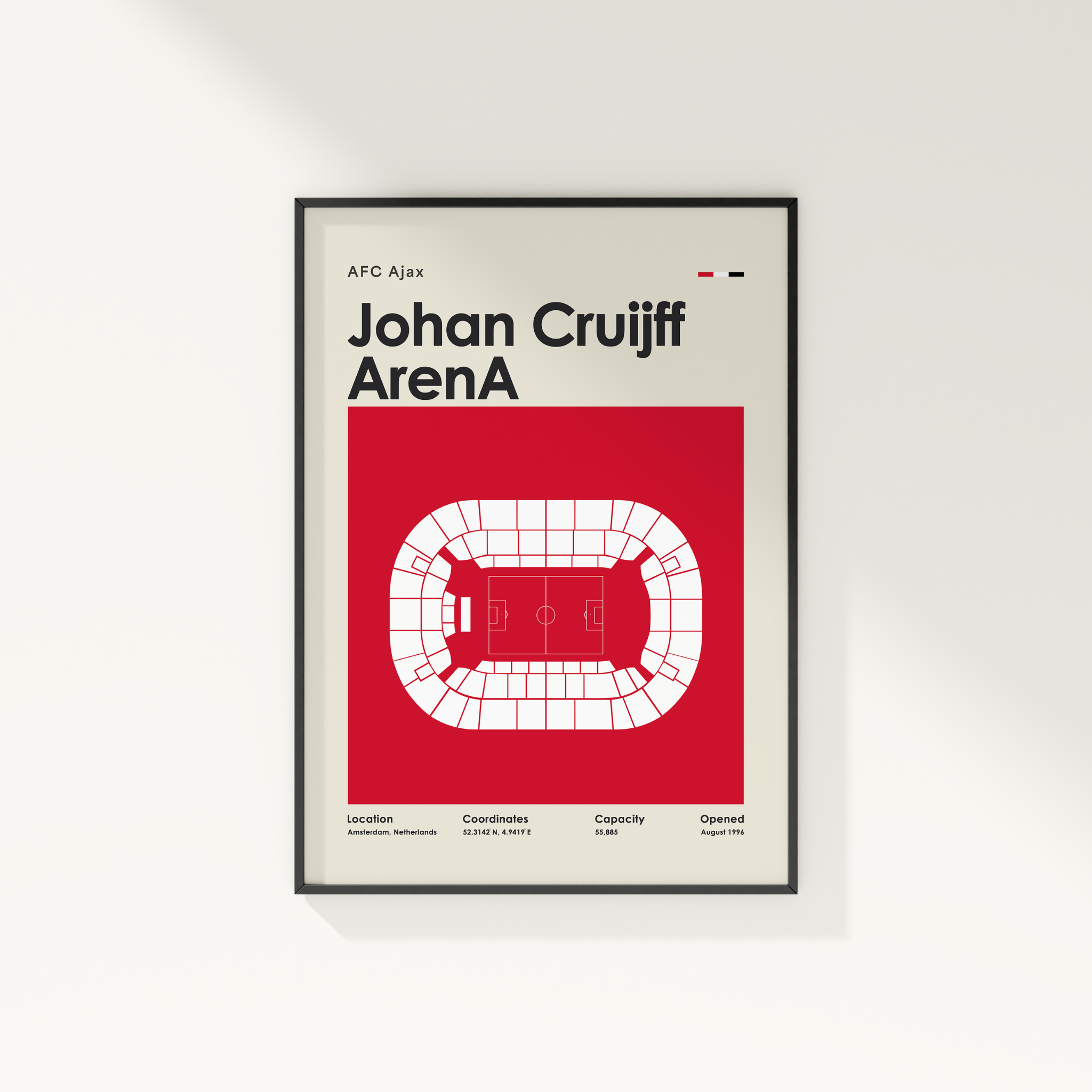 A framed minimalist sports poster of Johan Cruyff's Ajax stadium, featuring a bold red background, white stadium graphic, and black text on a light beige background, shown hanging on a white wall.