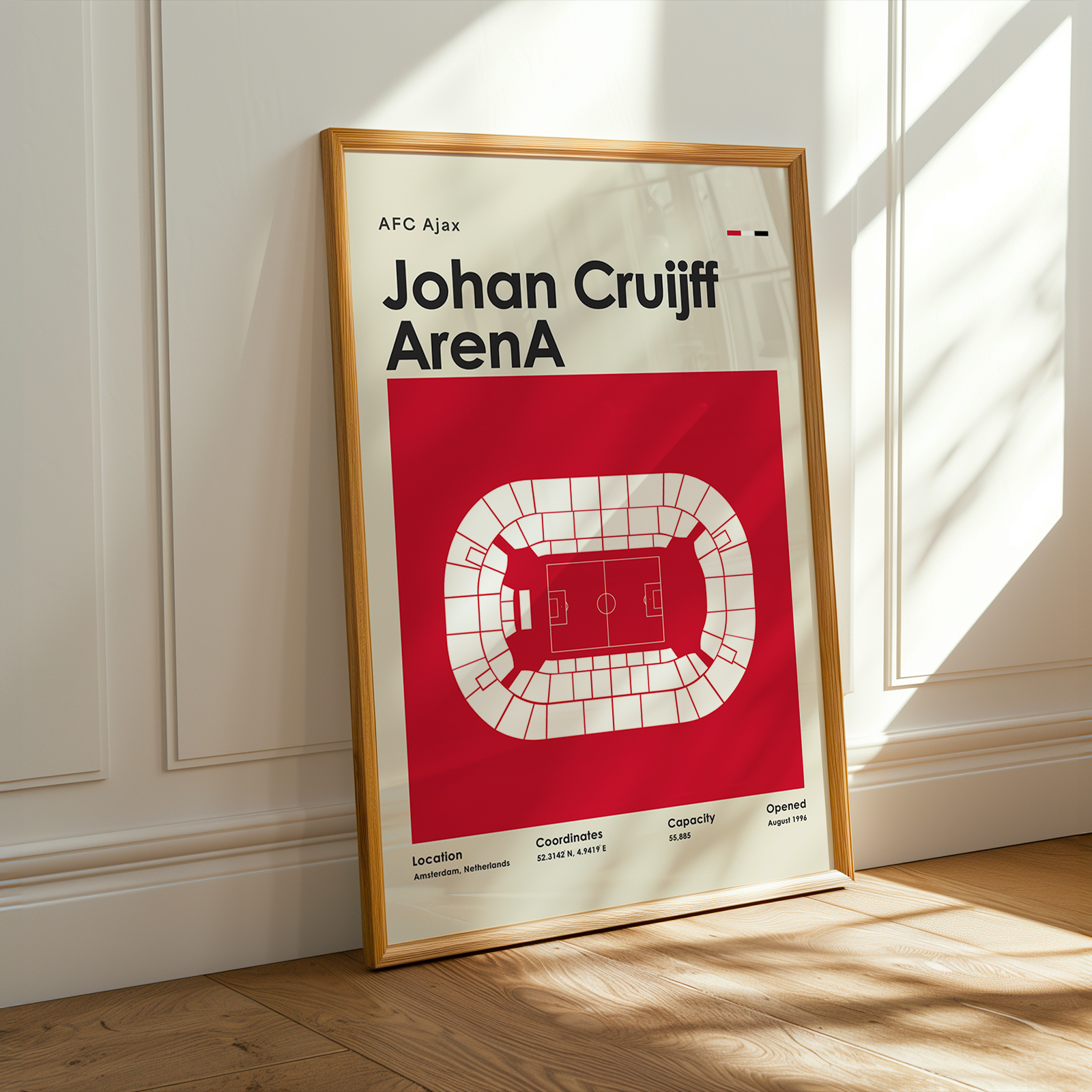 A framed sports poster of Johan Cruyff's Ajax stadium layout, leaning against a white paneled wall on a wooden floor with warm sunlight streaming in.