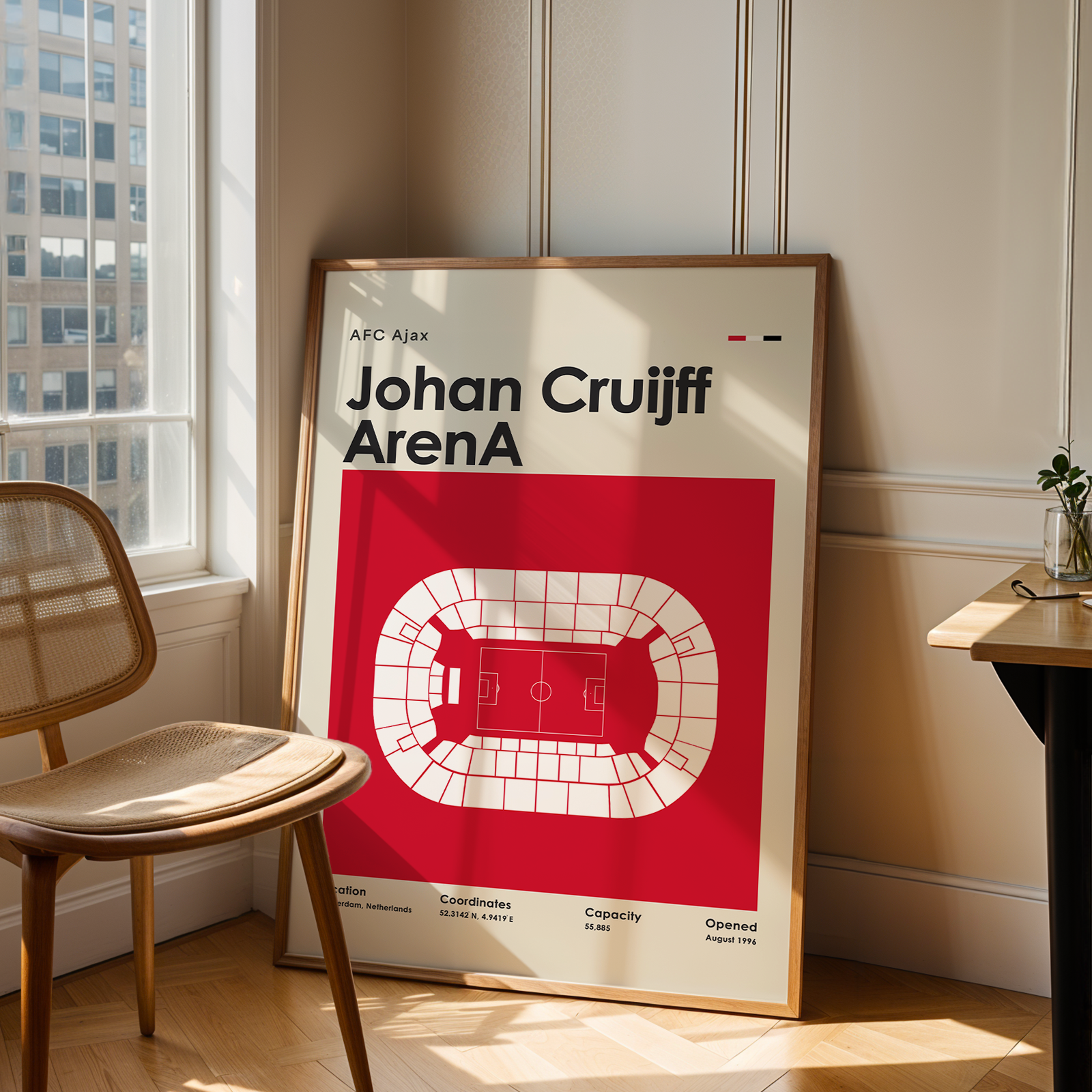 A framed sports poster of Johan Cruyff ArenaA from AFC Ajax leaning against a wall in a modern room with a wooden chair and desk.