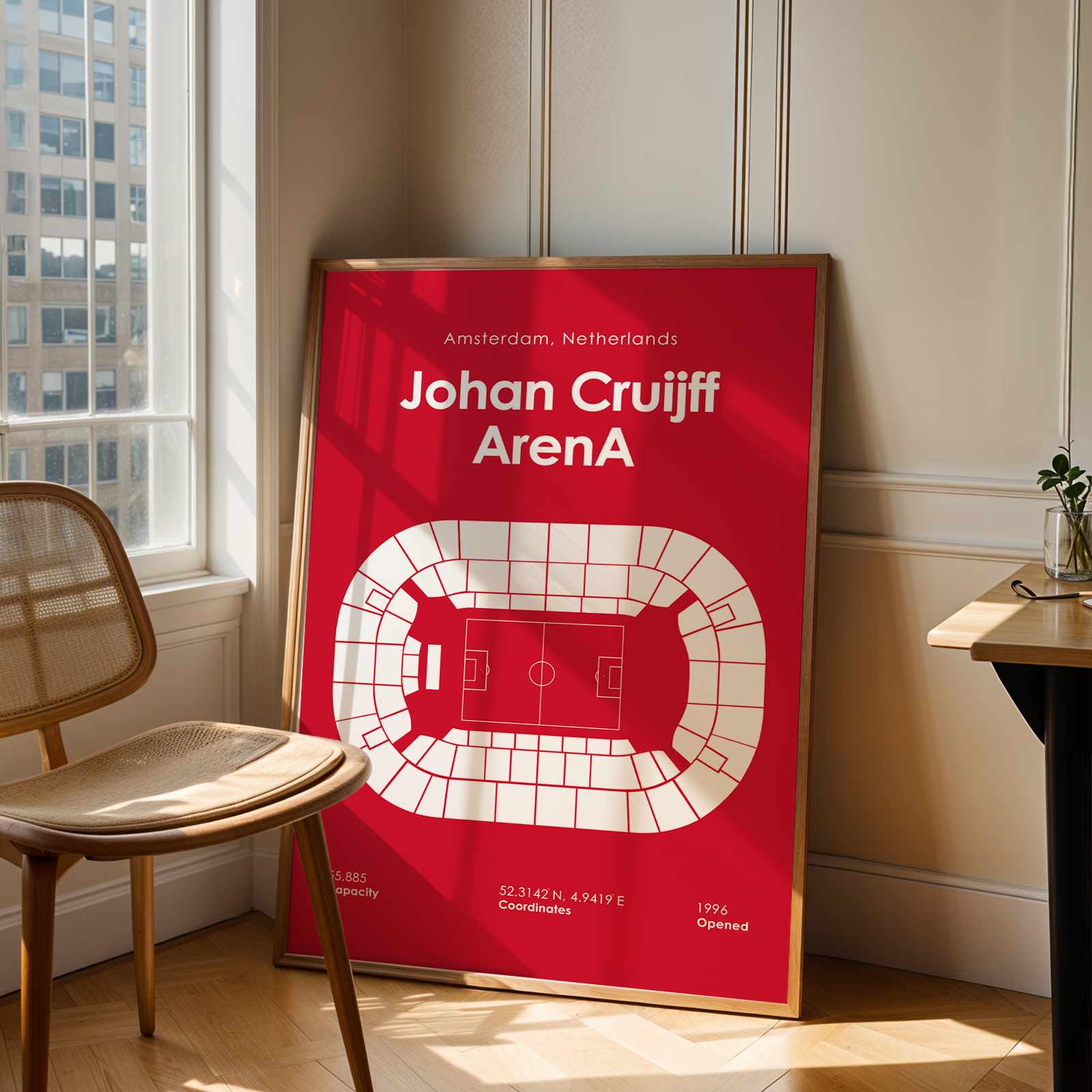 A framed red and white football stadium poster featuring Johan Cruijff and ArenA, leaning against a wall in a sunlit room next to a chair and desk.