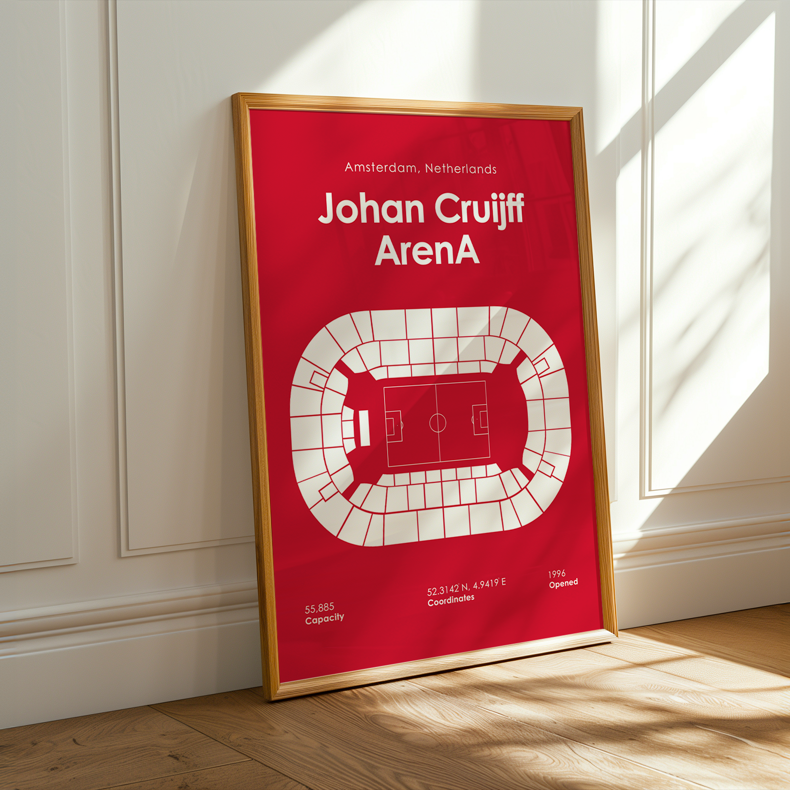 A framed sports poster of Johan Cruyff's Amsterdam, Netherlands, Arena stadium, leaning against a white paneled wall on a wooden floor with warm sunlight streaming in from a window.