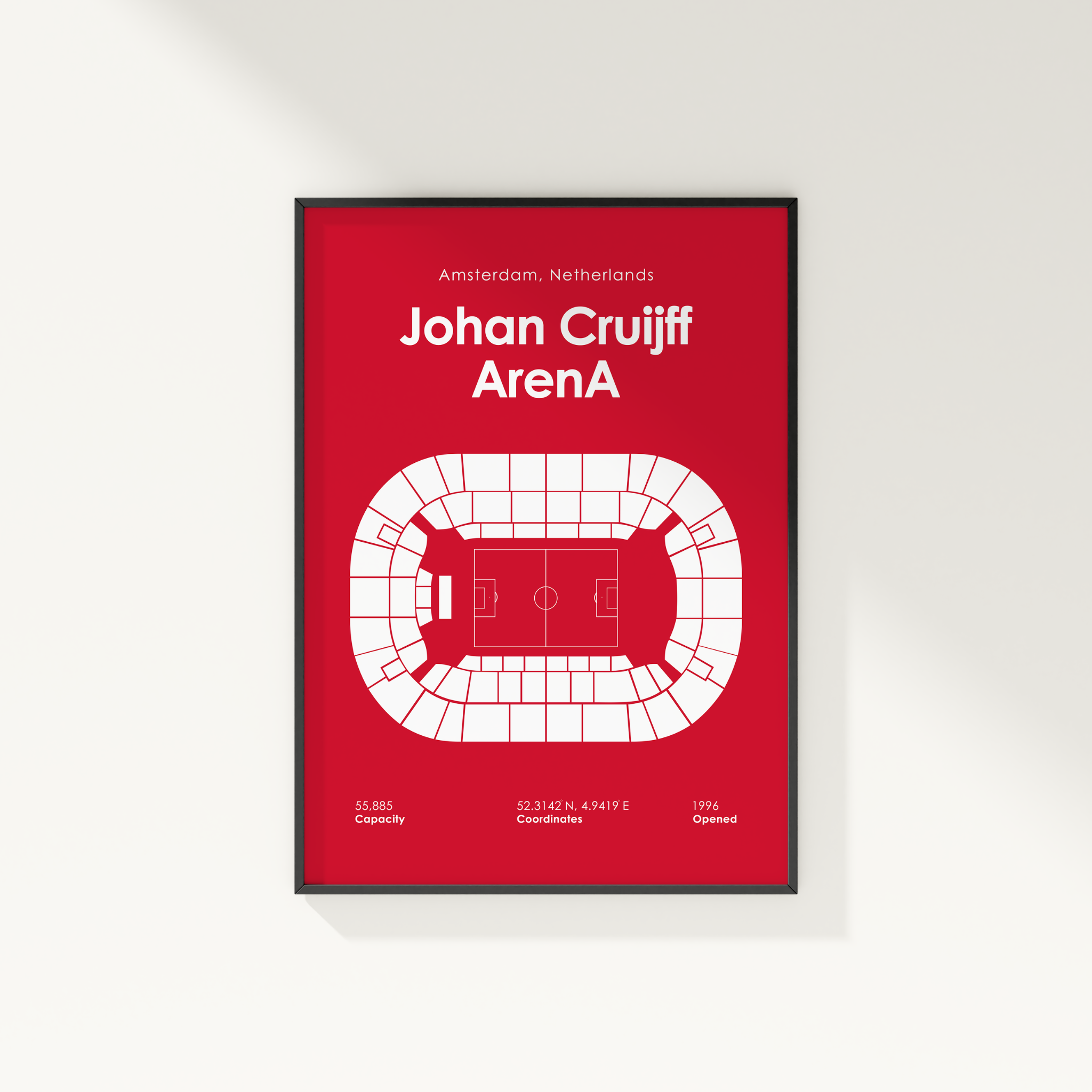 A framed minimalist poster featuring a white stadium floor plan and text reading 'Johan Cruijff ArenaA' on a red background, mounted on a light gray wall.
