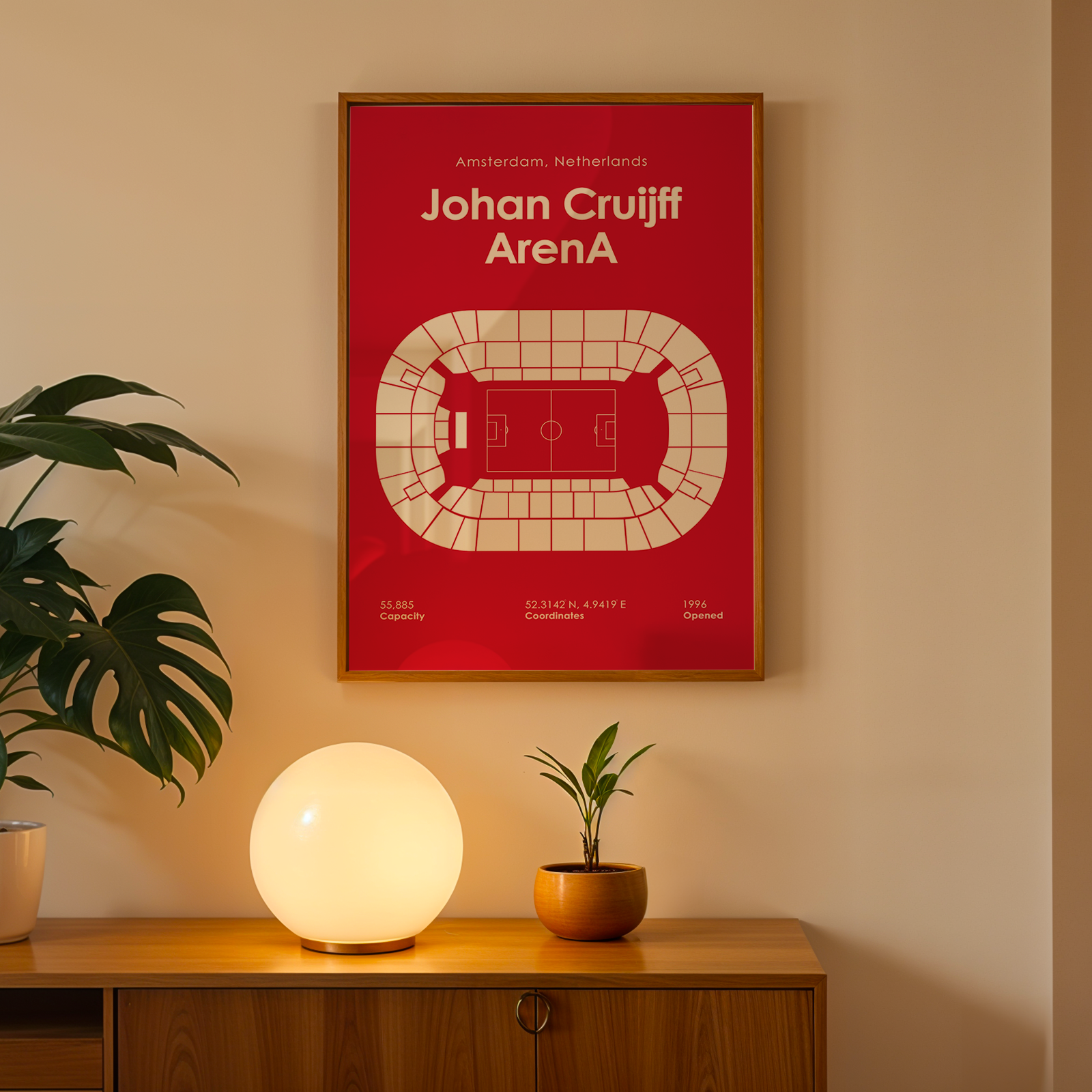 A framed red poster of Johan Cruyff Arena hanging on a beige wall above a wooden console table with a glowing globe lamp and potted plants.