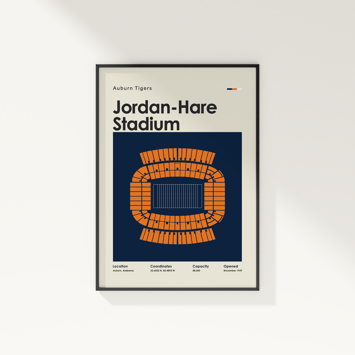 A framed poster print of Auburn Tigers Jordan-Hare Stadium, featuring a stylized orange stadium layout on a navy blue background with a light beige outer border, shown hanging on a white wall.