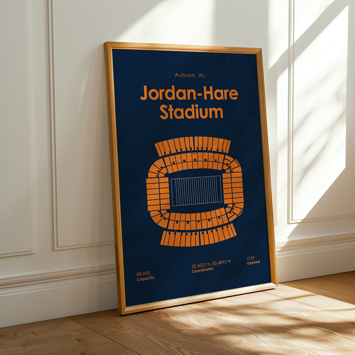A framed vertical poster leaning against a white paneled wall on a light wood floor, with warm sunlight and window shadows across the scene. The poster reads 'Jordan-Hare Stadium' with a blue background and orange stadium diagram.