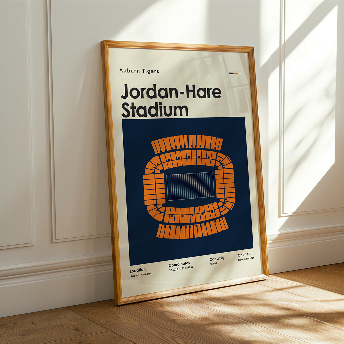 A framed poster of Jordan-Hare Stadium from the Auburn Tigers, leaning against a white paneled wall on a wooden floor with warm sunlight streaming in from the side.