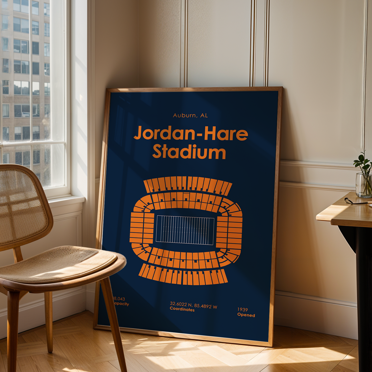 A framed poster of Jordan-Hare Stadium in Auburn, AL, displayed on a wooden floor in a bright room next to a chair and a desk.